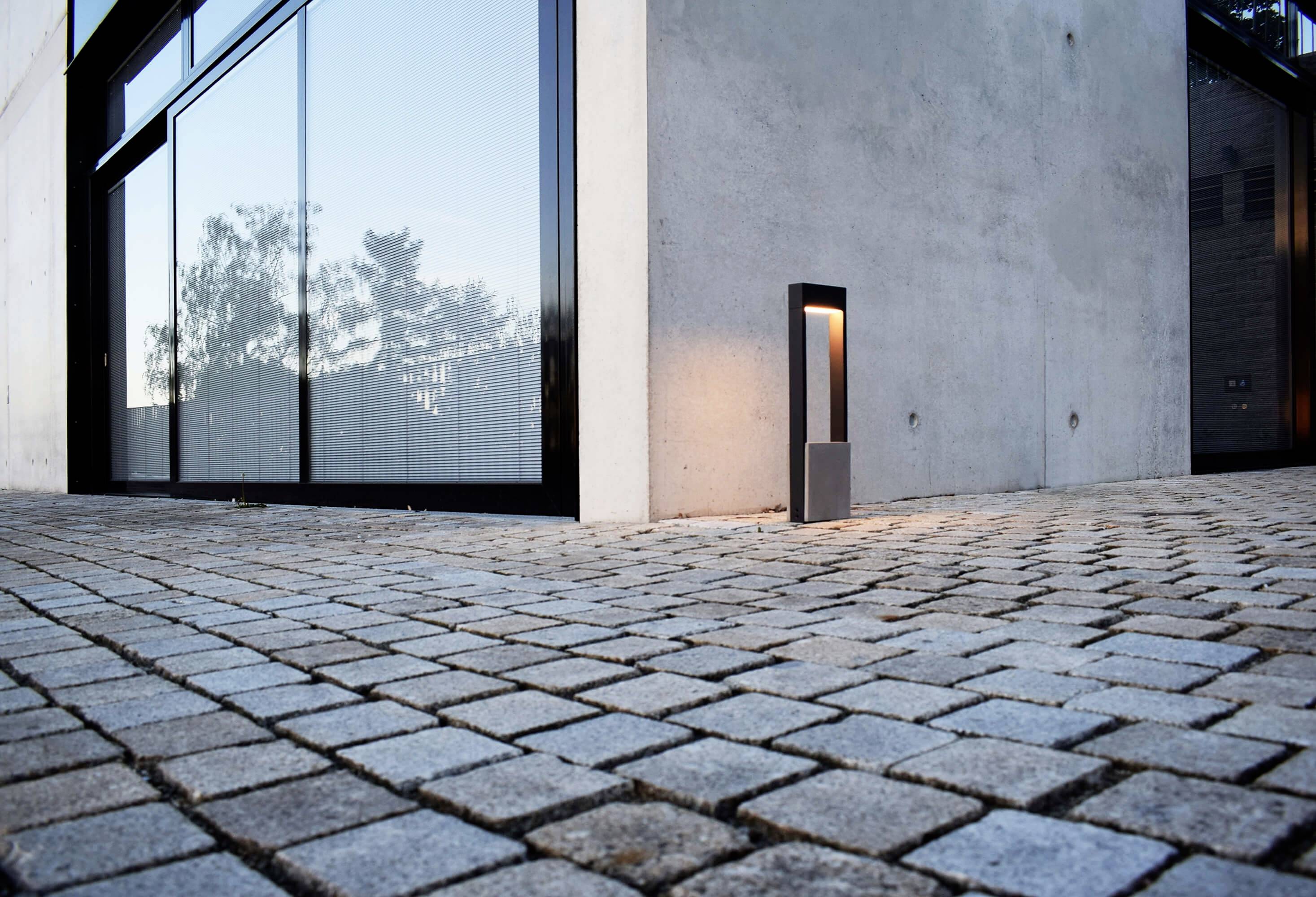 Ein modernes Gebäude mit großen Glasfenstern, umgeben von einem gepflasterten Boden. Eine Außenleuchte steht neben der Wand.