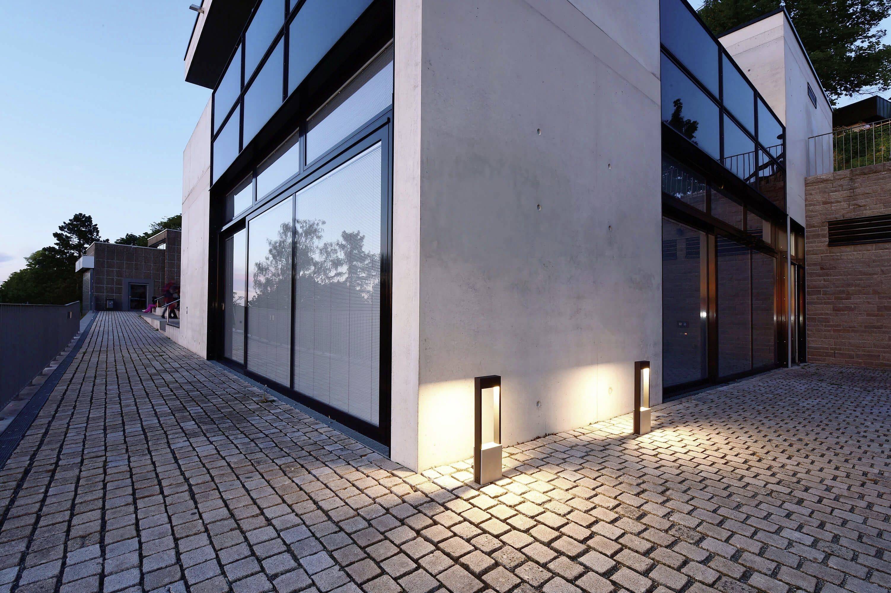 Modernes Gebäude mit großen Fenstern, umgeben von Pflastersteinen. Abendszene mit eingeschalteter Außenbeleuchtung.