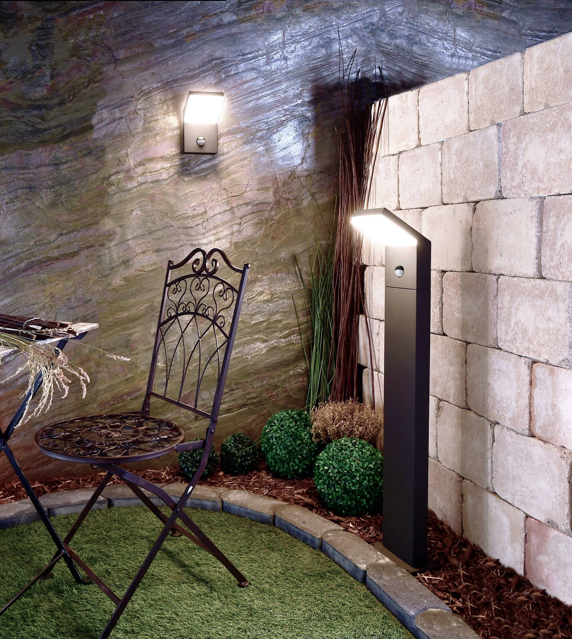 Ein Sitzbereich im Freien mit schwarzem Metallstuhl, künstlichem Rasen und dekorativen Pflanzen, beleuchtet von zwei modernen Wandlampen.