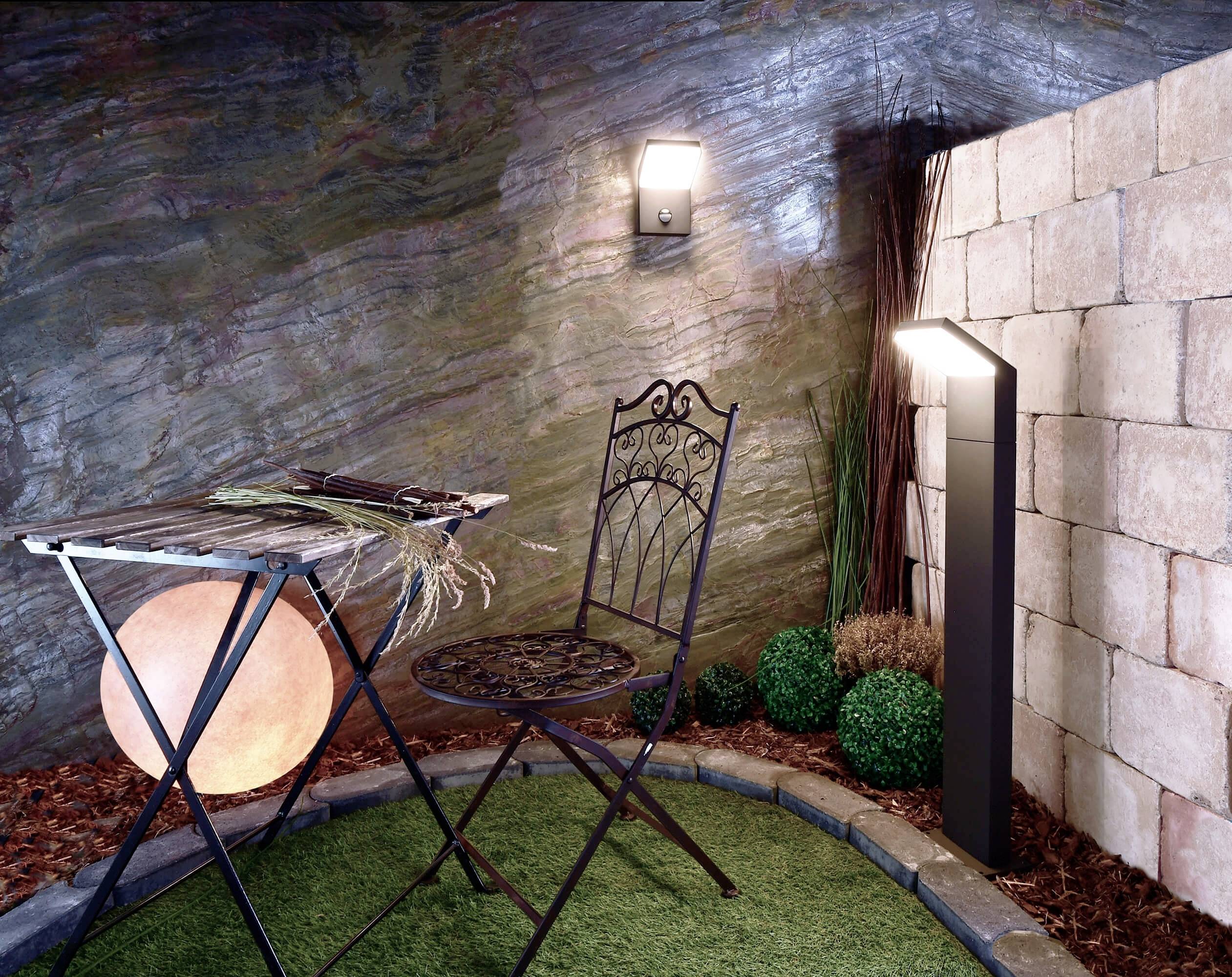 Ein kleiner Gartenbereich mit einem schmiedeeisernen Tisch und Stuhl auf Kunstrasen. Eine Wandlampe und Leuchte erhellen die Szene.