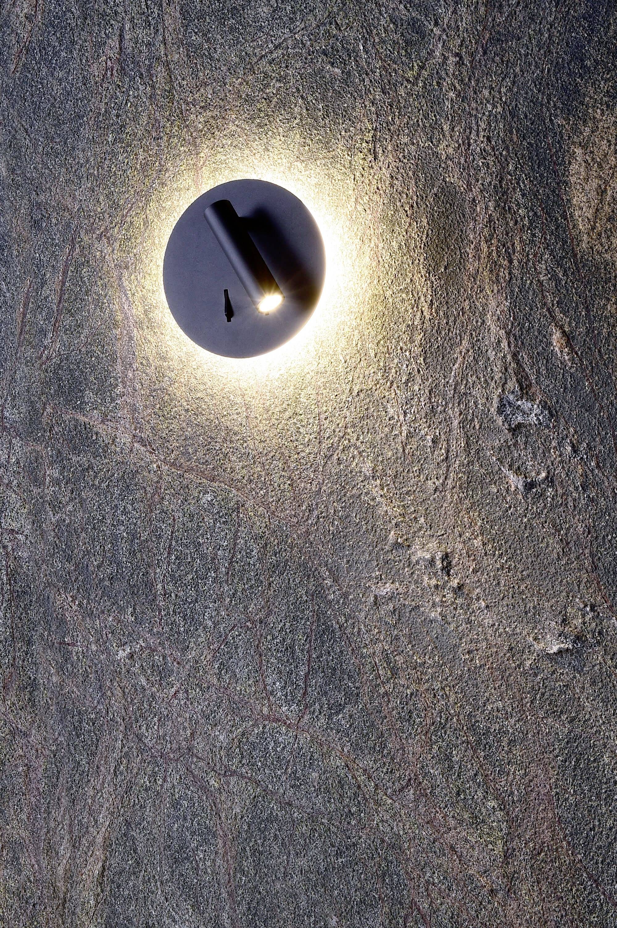Leuchte in runder Form, die warmes Licht auf eine strukturierte Steinwand strahlt.
