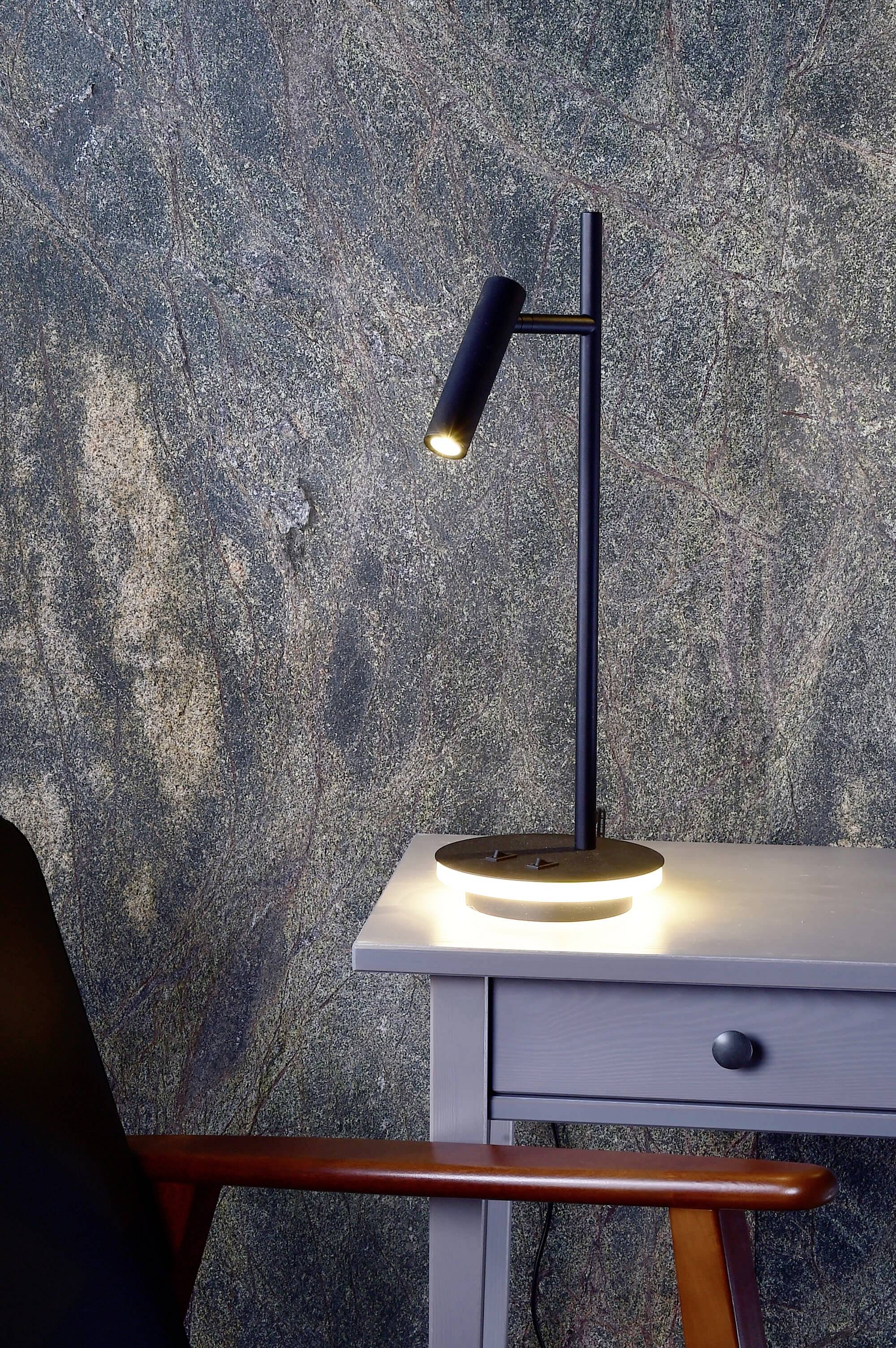 Eine schlanke, schwarze Tischlampe leuchtet auf einem grauen Holztisch vor einer strukturierten Wand. Daneben steht ein Stuhl.