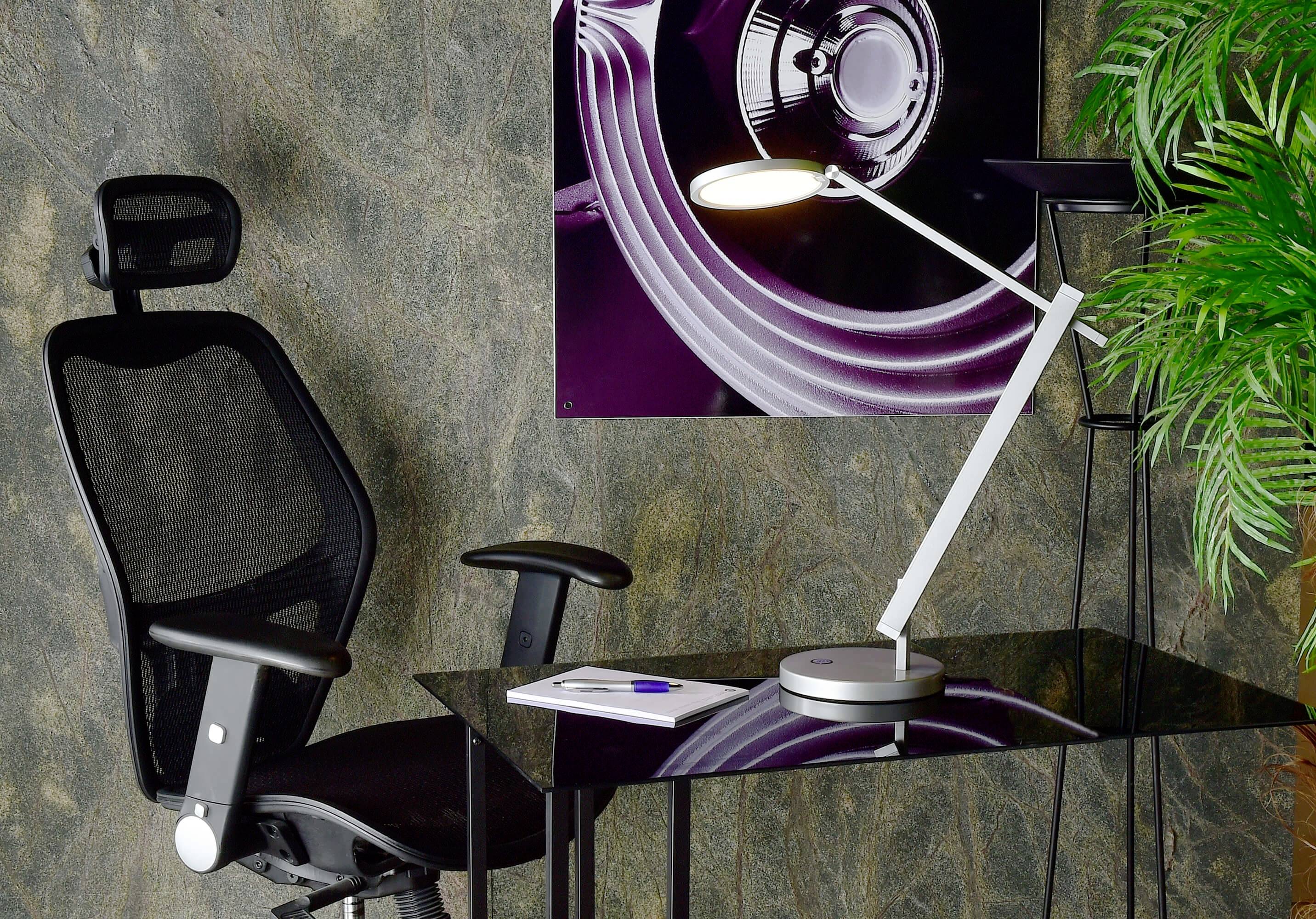 Ein moderner Bürostuhl und Schreibtisch mit Lampe und Notizbuch, vor einer Wand mit abstraktem Kunstwerk und Pflanze.