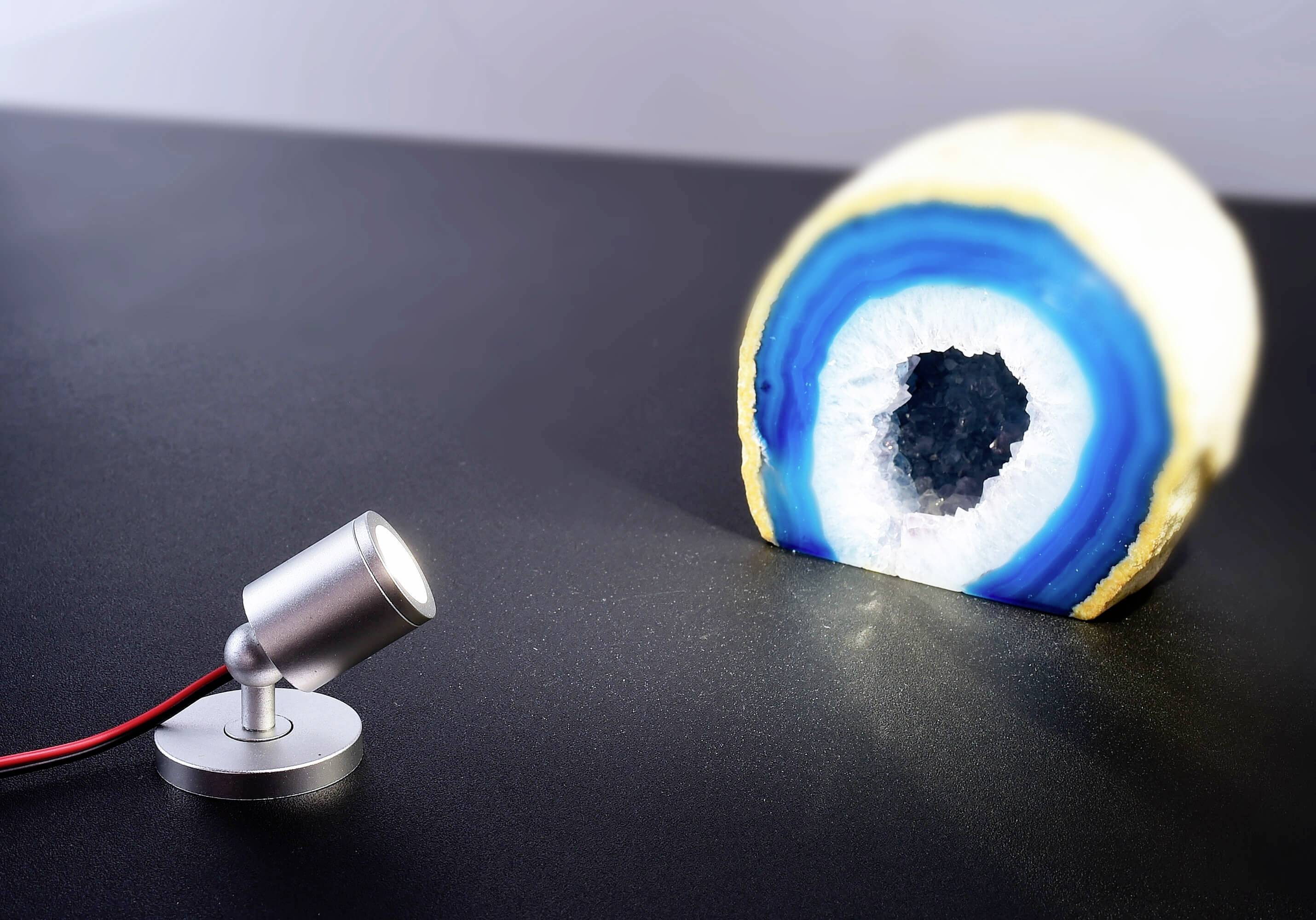 Ein leuchtender Lampenspot beleuchtet eine polierte, halbierte blau-weiße Geode auf einem schwarzen Tisch und zeigt deren innere Kristalle.