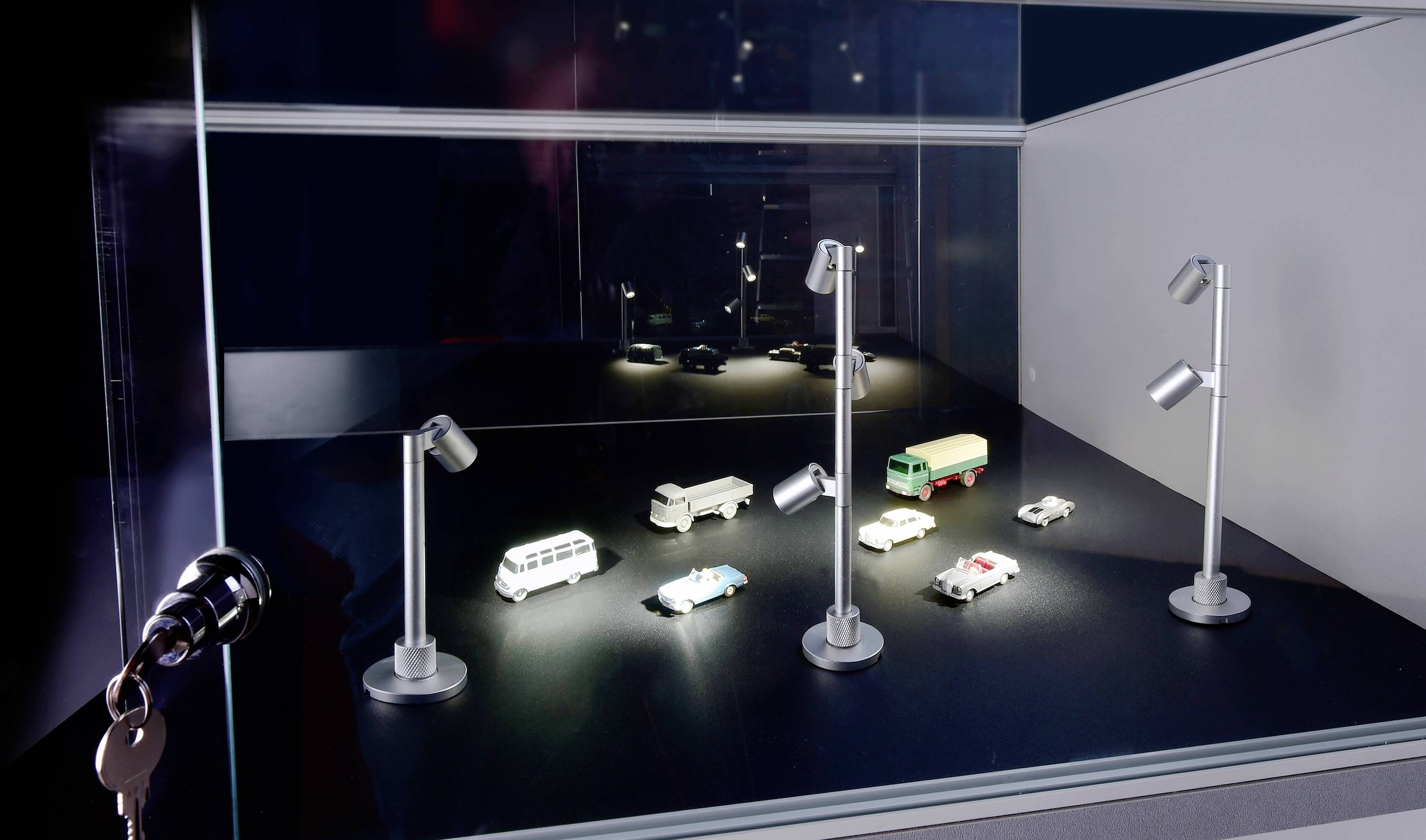 Miniatur-Szene mit mehreren beleuchteten Modellfahrzeugen auf einer schwarzen Fläche, umgeben von kleinen Straßenlaternen in einem Schaukasten.