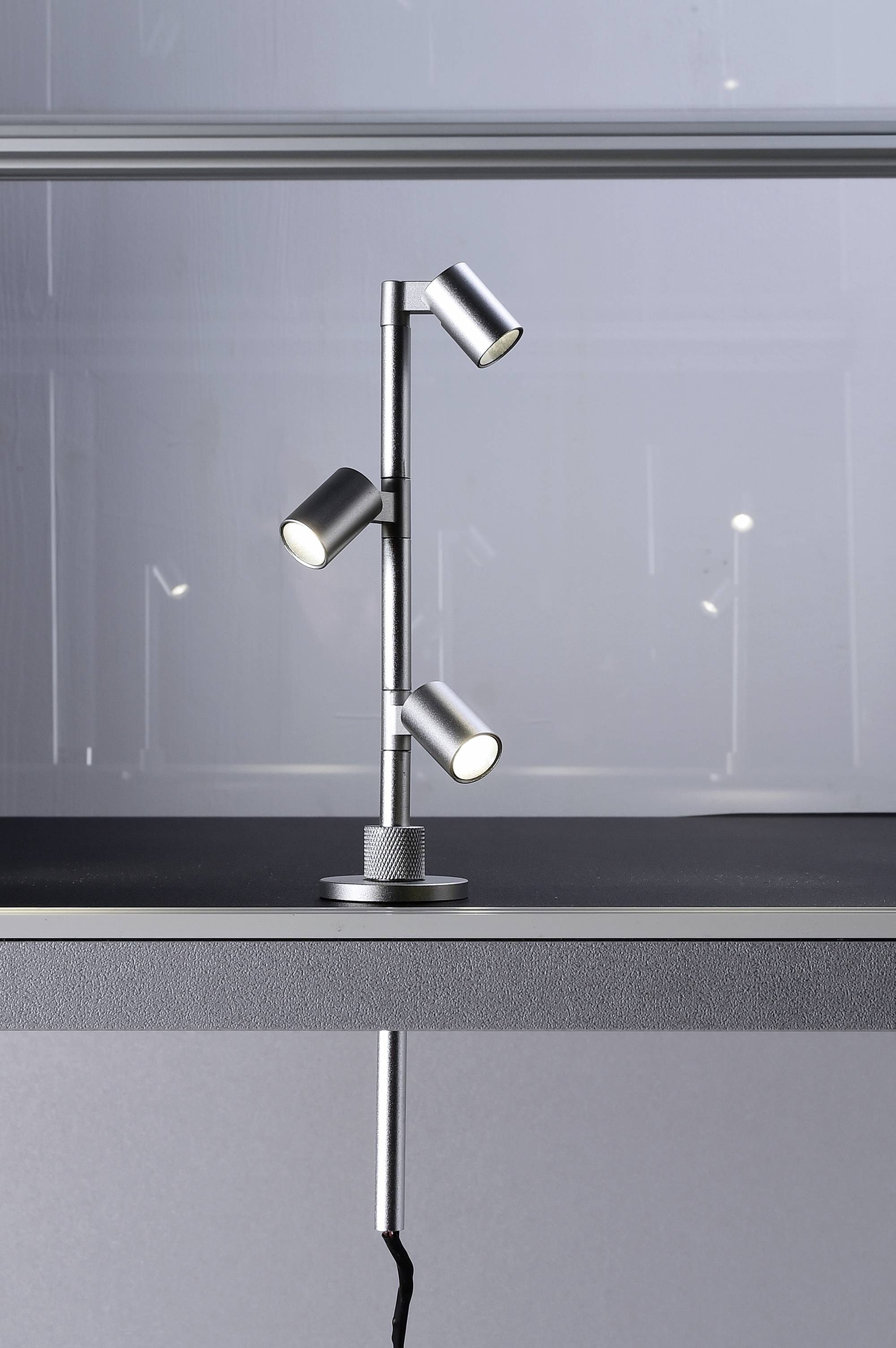 Eine modern gestaltete Tischlampe mit drei verstellbaren Leuchten auf einem Metallgestell, platziert auf einem grauen Tisch.