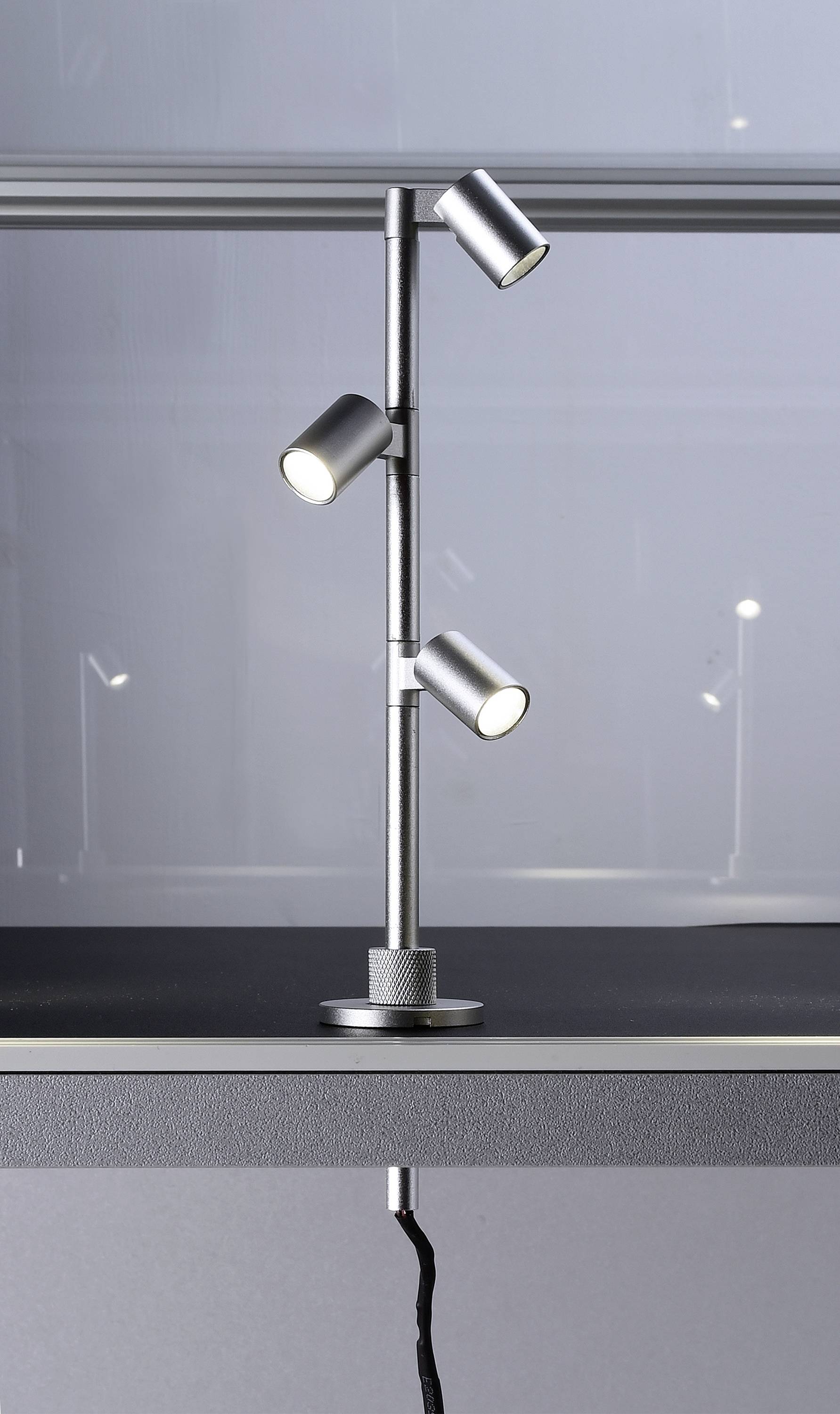 Silberne, dreiköpfige Schreibtischlampe mit verstellbaren Spots auf einer grauen Oberfläche, indirekte Beleuchtung erzeugend.