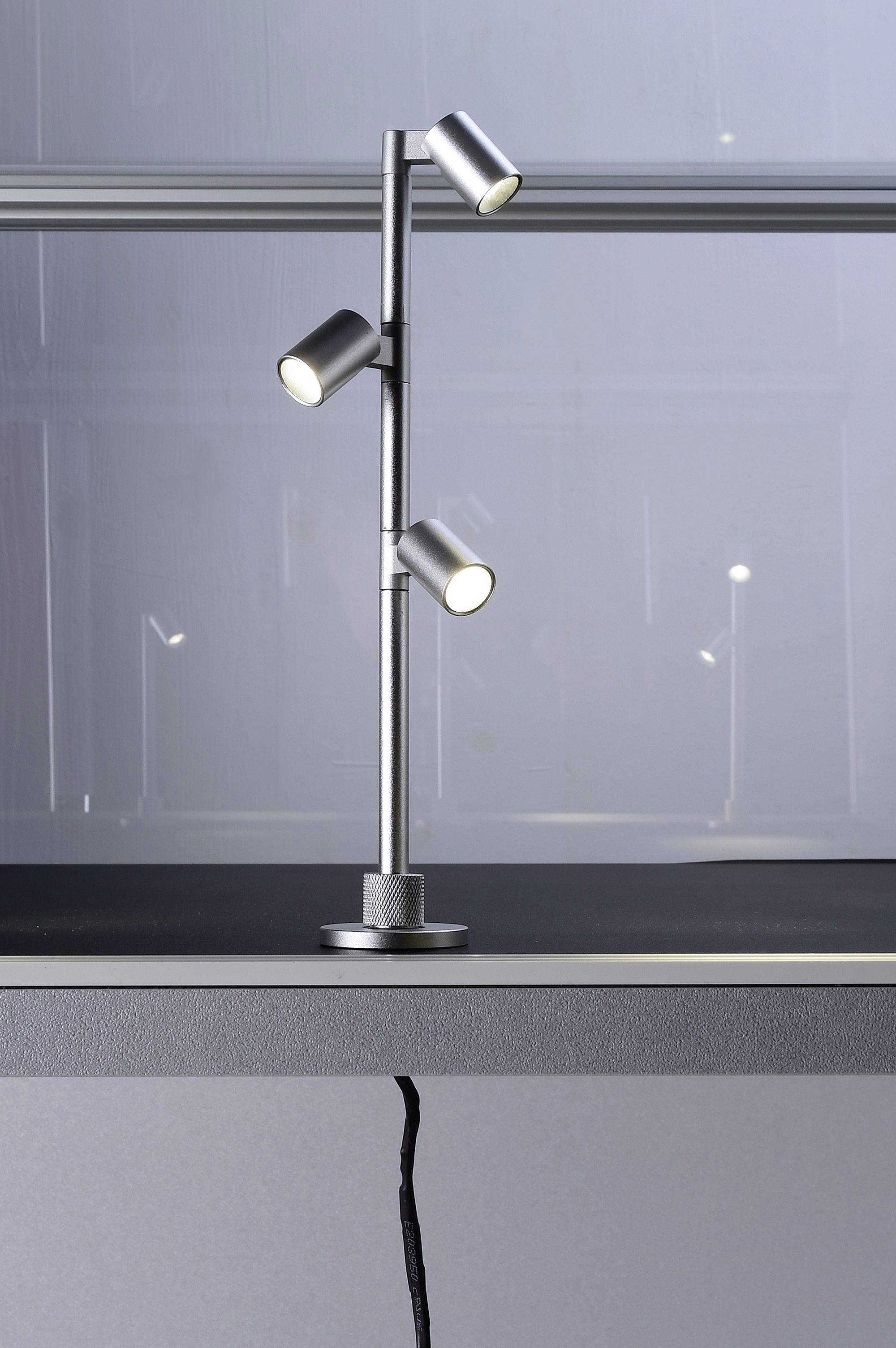 Eine moderne, silberne Tischlampe mit drei verstellbaren Leuchten auf einem Regal, die Umgebung ist minimalistisch gestaltet.