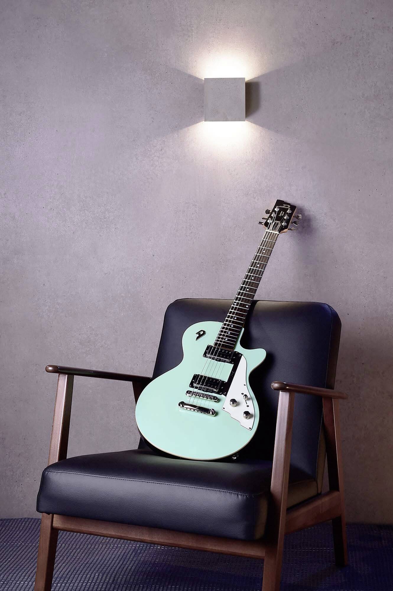 Eine elektrische Gitarre mit türkisem Korpus liegt auf einem schwarzen Ledersessel vor einer grauen Wand mit einer leuchtenden Wandlampe.