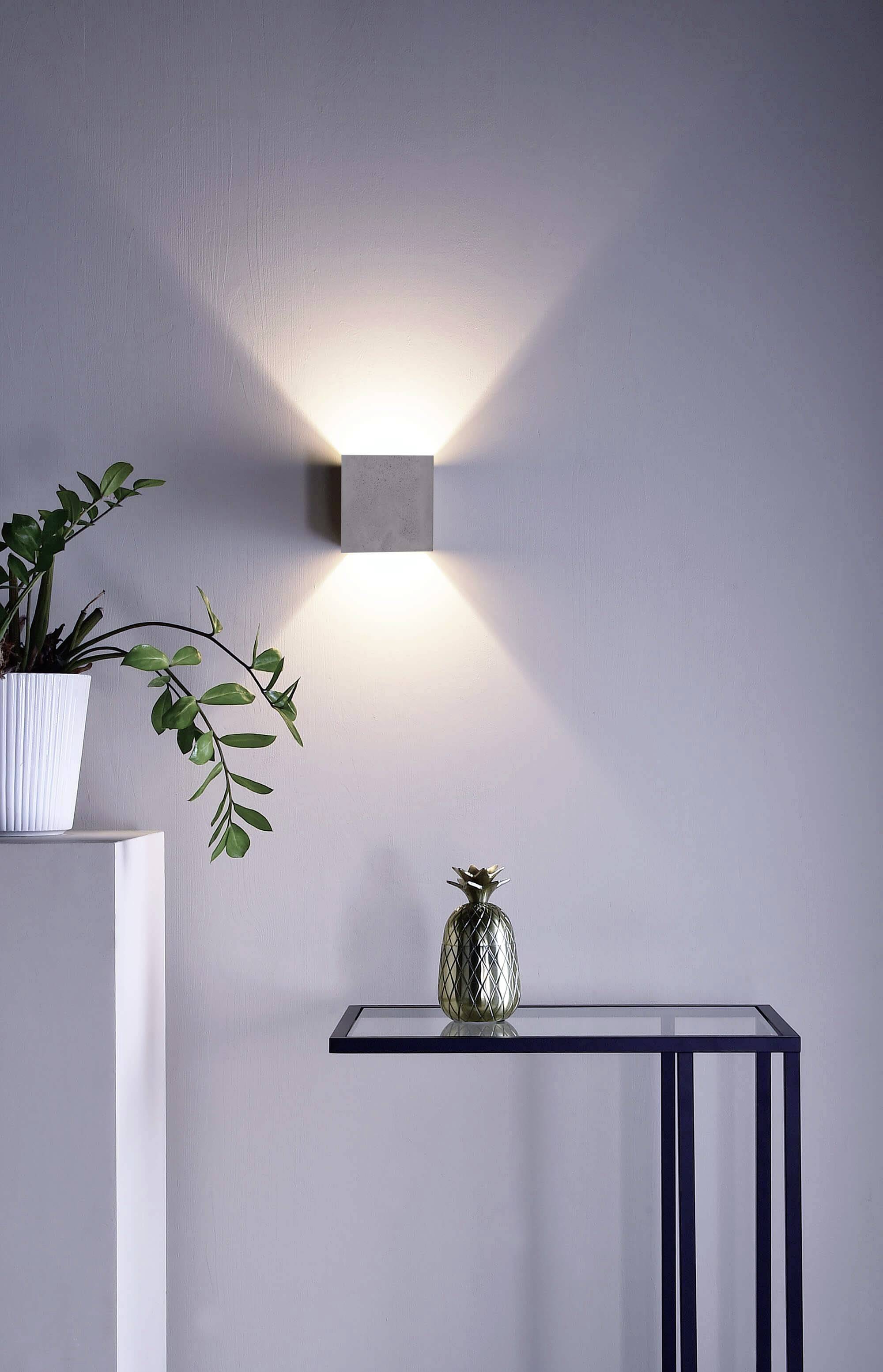 Wandleuchte strahlt warmes Licht aus. Daneben ein kleiner Tisch mit Glasoberfläche, darauf steht eine Ananas-Deko. Links steht eine Pflanze in einem weißen Topf.