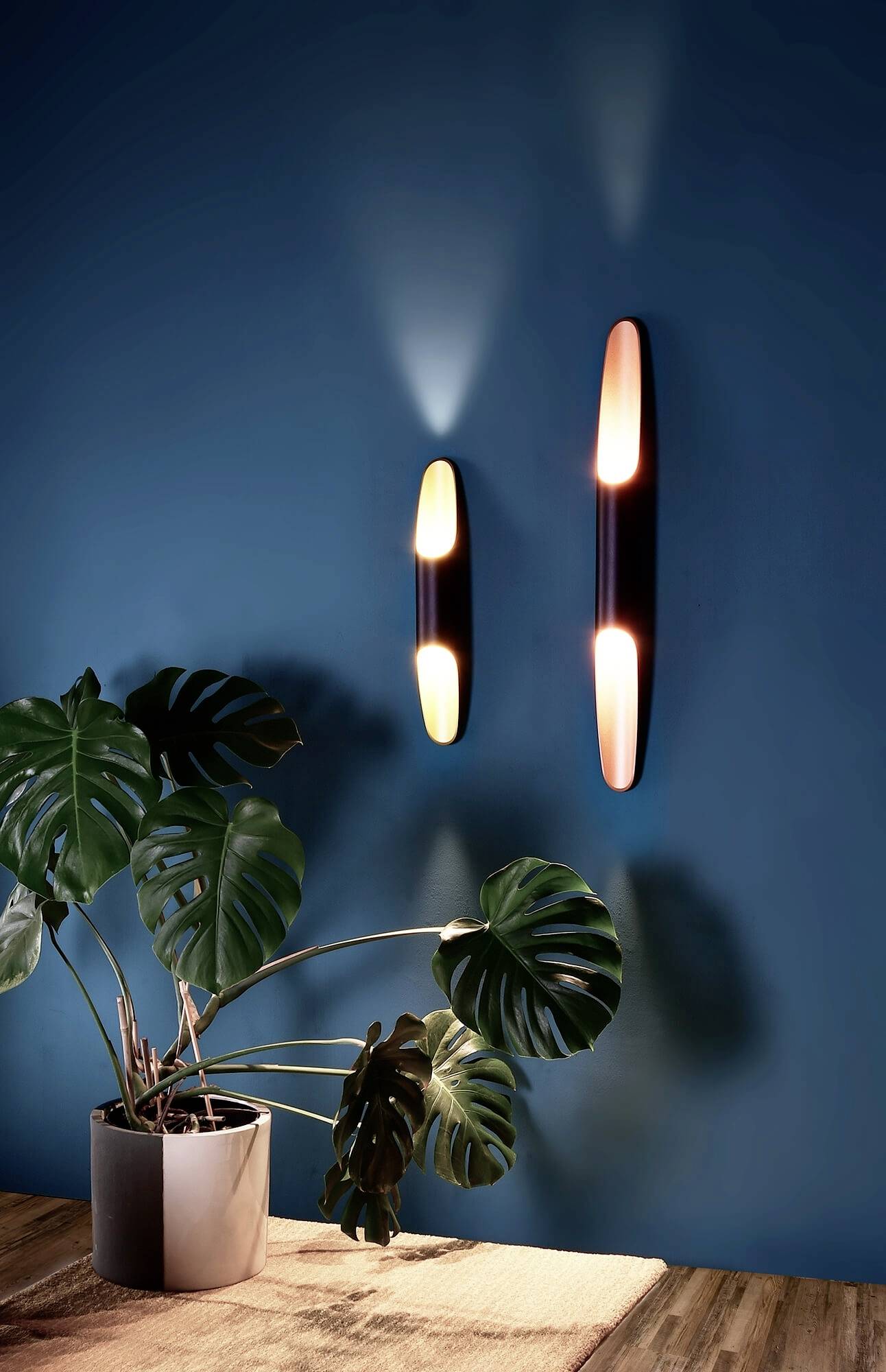 Zwei moderne Wandleuchten strahlen warmes Licht aus, montiert auf einer blauen Wand. Daneben ist eine Pflanze in einem Topf zu sehen.