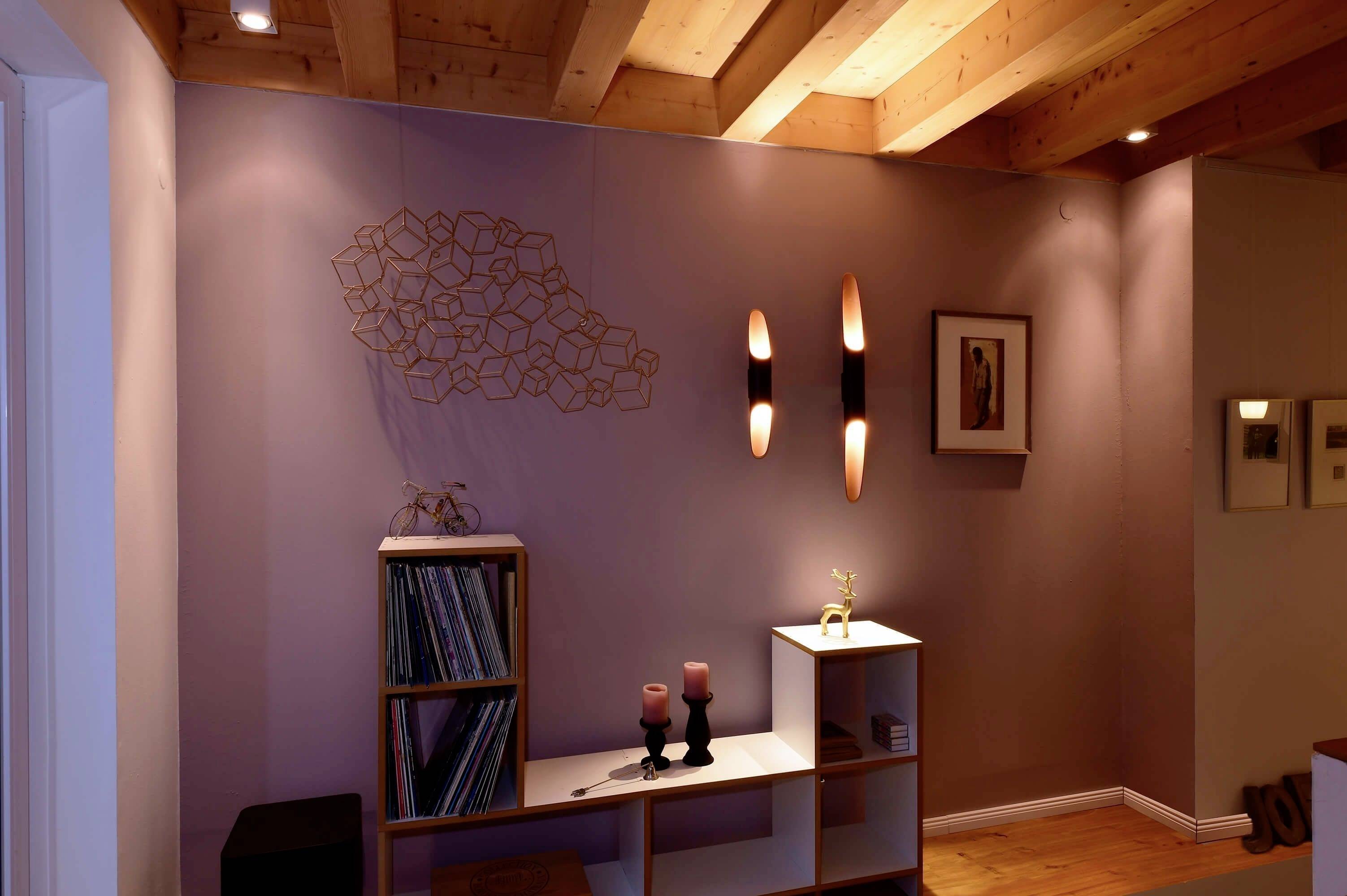 Ein Wohnzimmer mit Holzbalkendecke, einem Wandregal mit Deko-Kerzen und eingerahmten Bildern an der Wand. Gemütliche Beleuchtung.