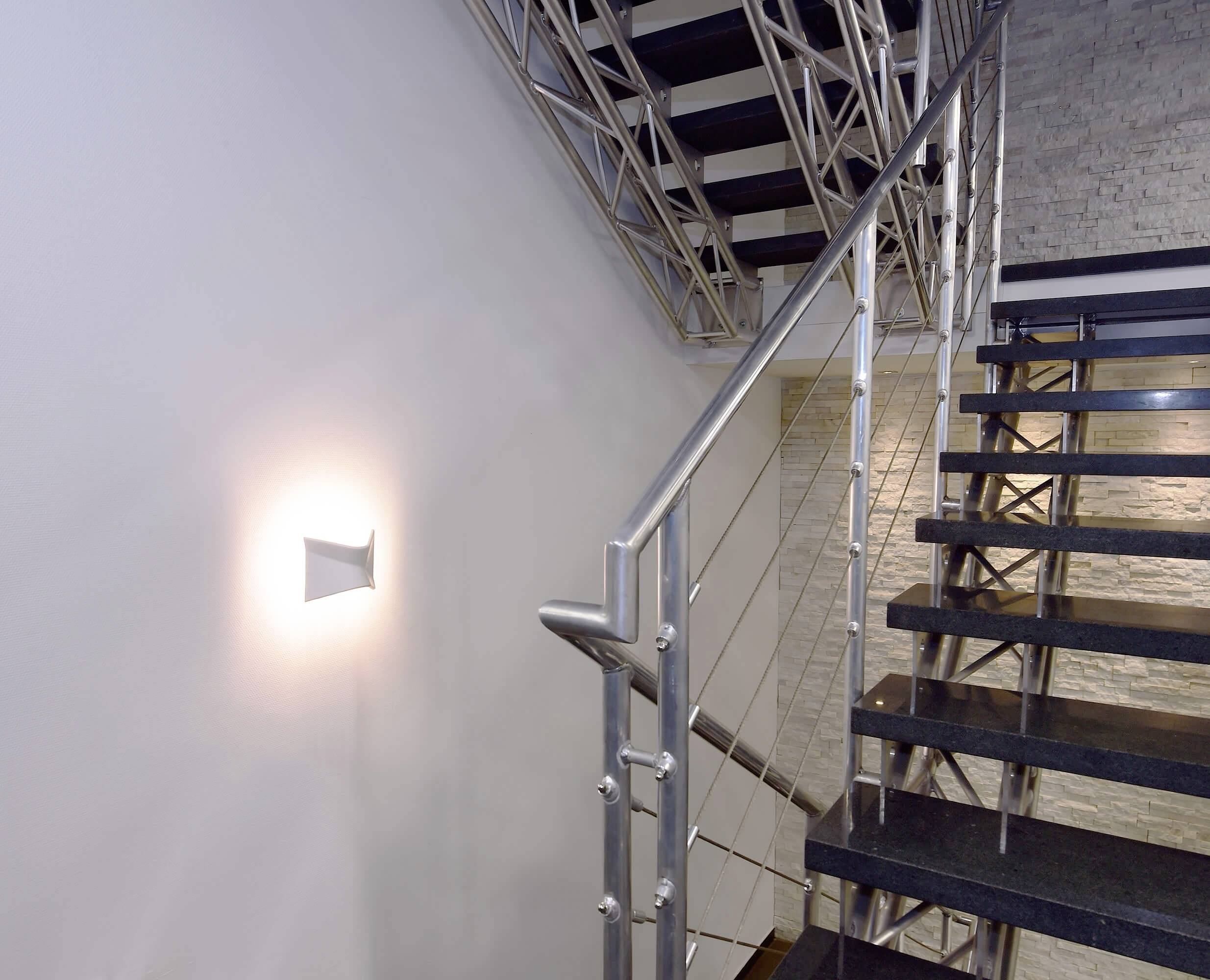 Eine moderne Treppe aus Metall und Holz mit einem minimalistisch beleuchteten weißen Wandlicht an der Seite. Treppenhaus in einem Gebäude.