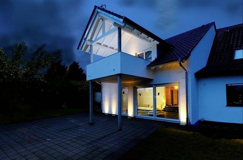 Ein modernes Einfamilienhaus bei Nacht, mit beleuchtetem Balkon und großen Fensterfronten, umgeben von einem gepflasterten Hof.
