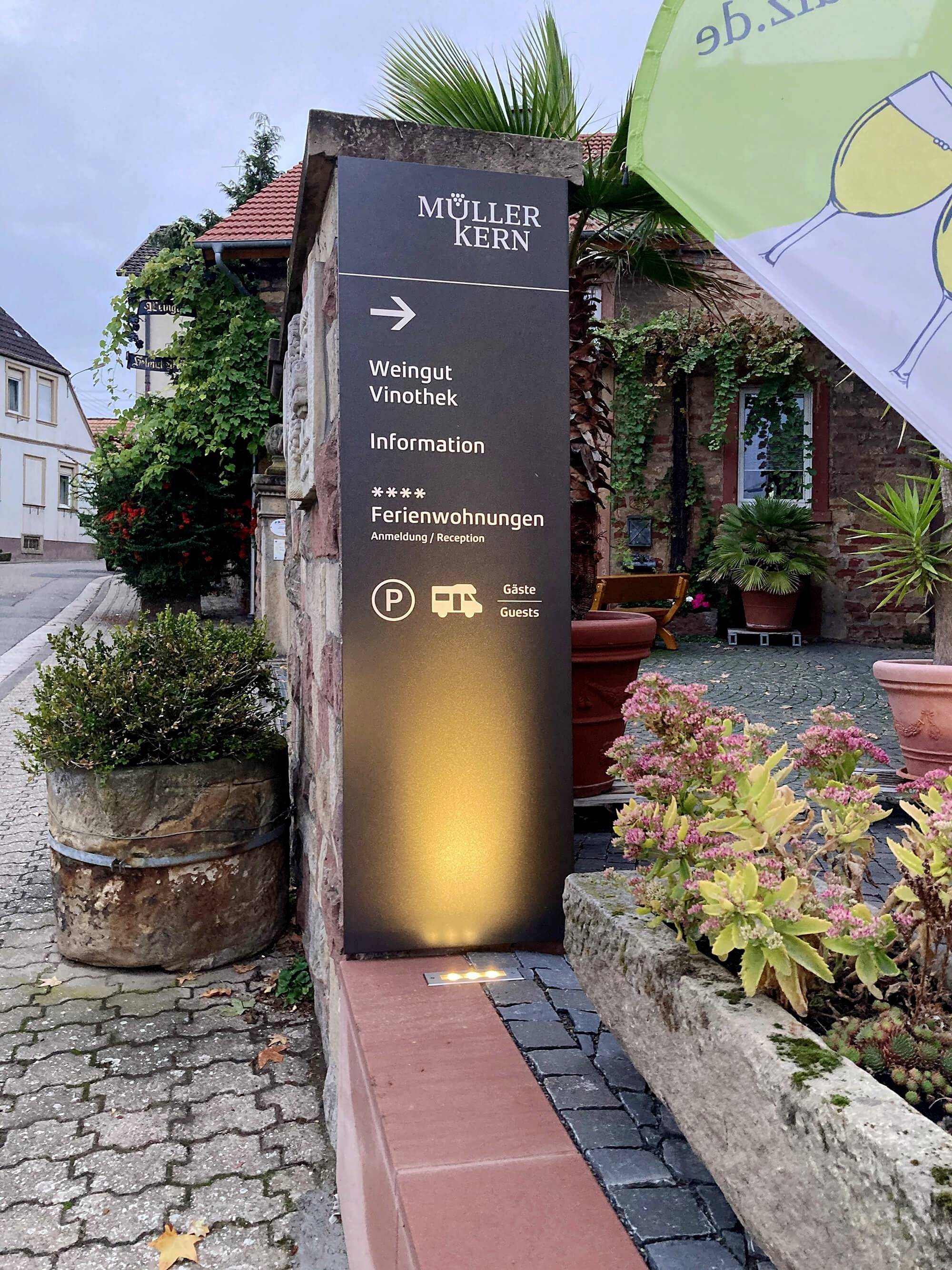 Wegeleitstein mit Aufschrift 'Müller Kern', weist auf Weingut, Vinothek und Ferienwohnungen hin. Umgebung zeigt Pflanzen und Gebäude.