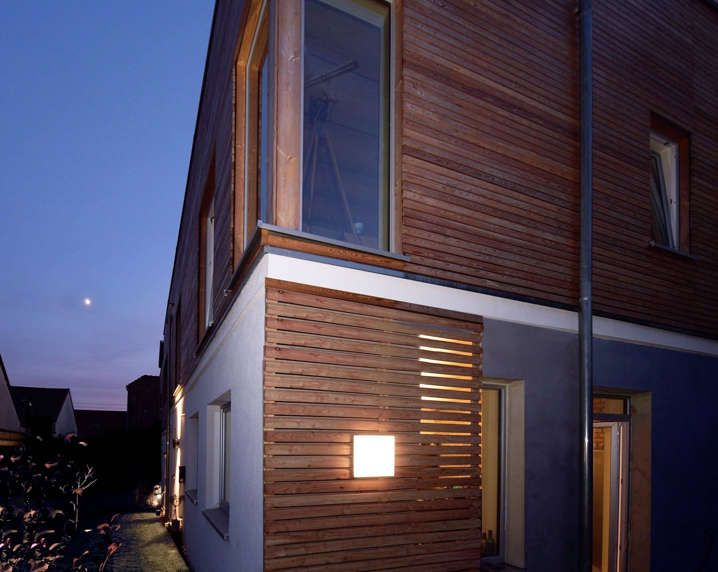 Eckansicht eines modernen Hauses bei Dämmerung, mit Holzverkleidung und beleuchtetem Fenster. Der Himmel ist leicht dunkelblau.