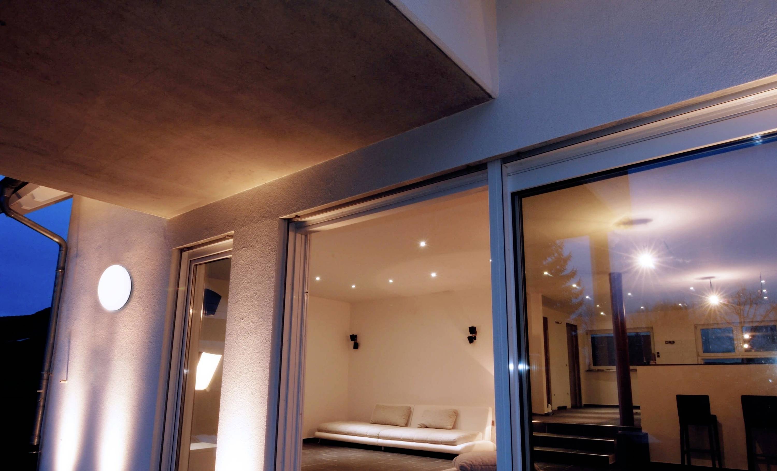 Großes Wohnzimmer mit moderner Beleuchtung, sichtbarer Couch und großen Glasfenstern, die eine Terrasse bei Dämmerung zeigen.