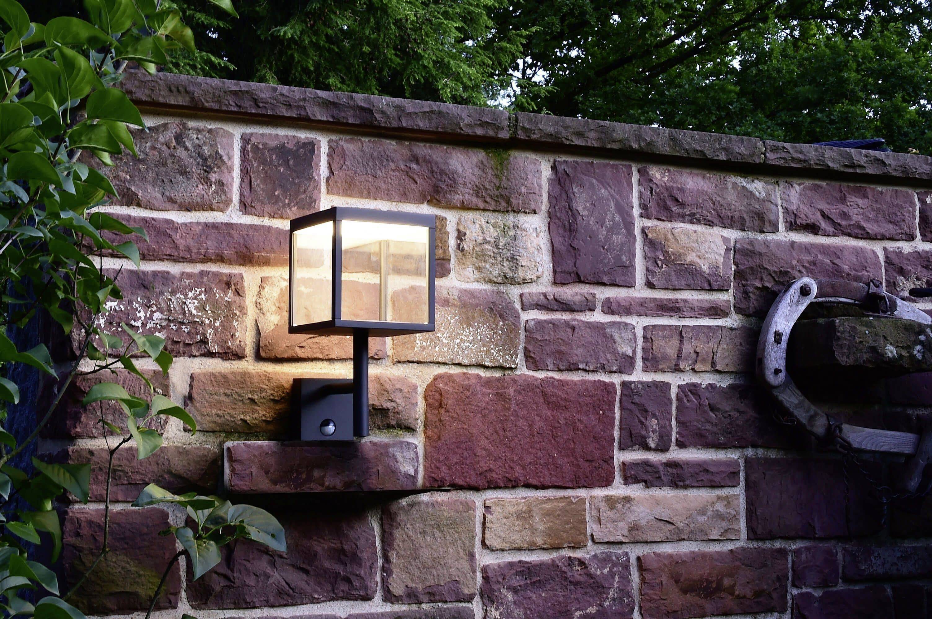 Eine Wandleuchte leuchtet auf einer rustikalen Steinmauer, umgeben von Pflanzen. Die Szene vermittelt eine gemütliche Gartenatmosphäre.