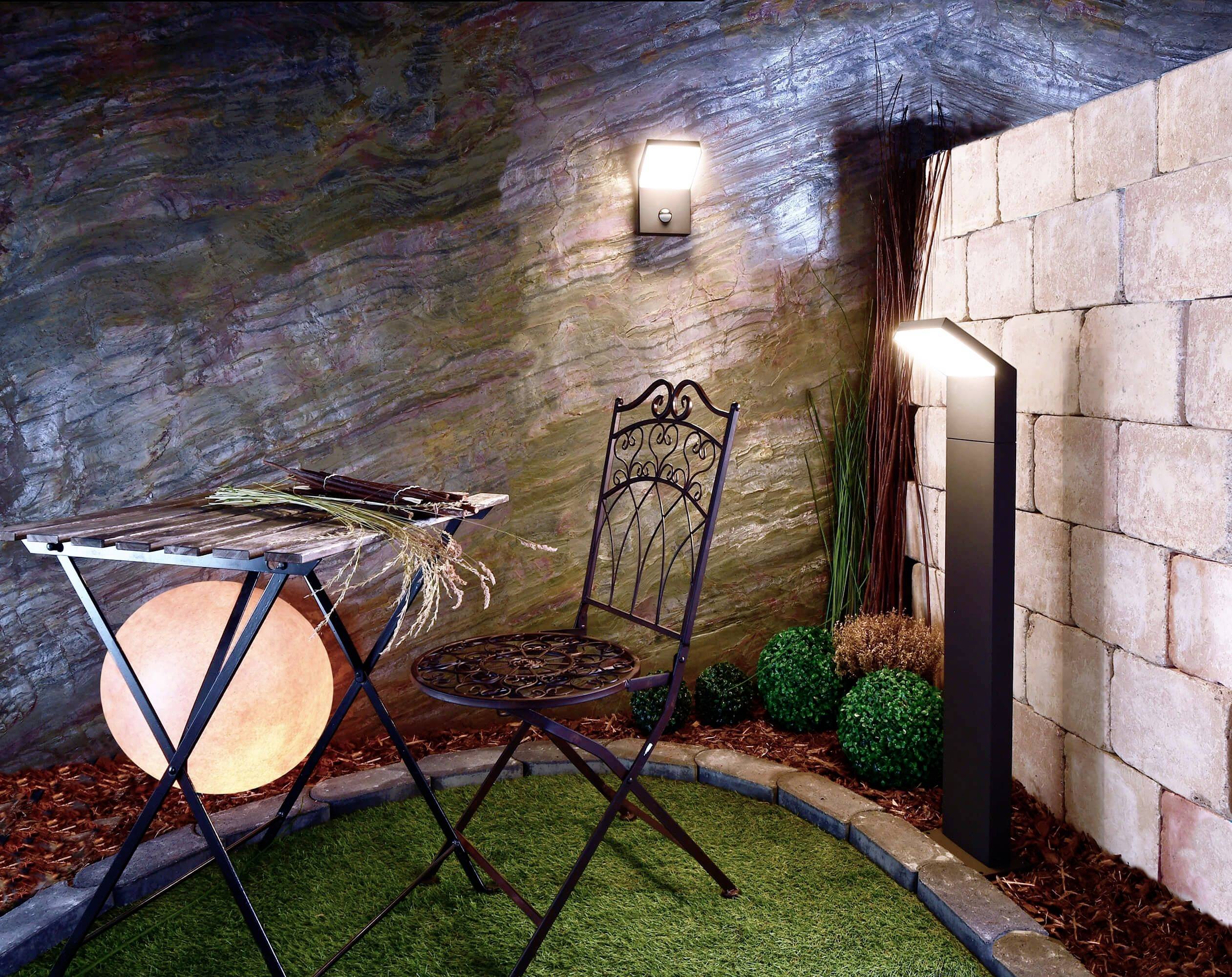 Gemütlicher Innenhof mit einem Metallstuhl und einem Tisch auf Kunstrasen, umgeben von Zierpflanzen und beleuchtet durch Wandlampen.