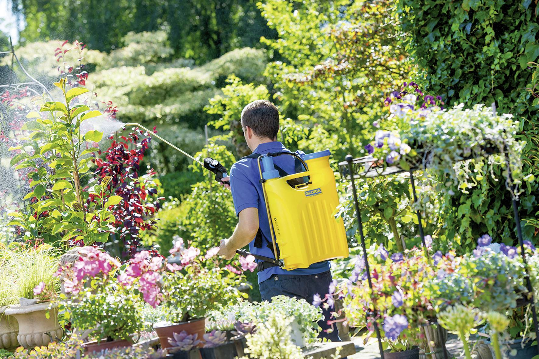 Ein Mann mit einem gelben Rucksack-Sprühgerät gießt Pflanzen in einem blühenden Garten. Bunte Blumen und grünes Laub umgeben ihn.