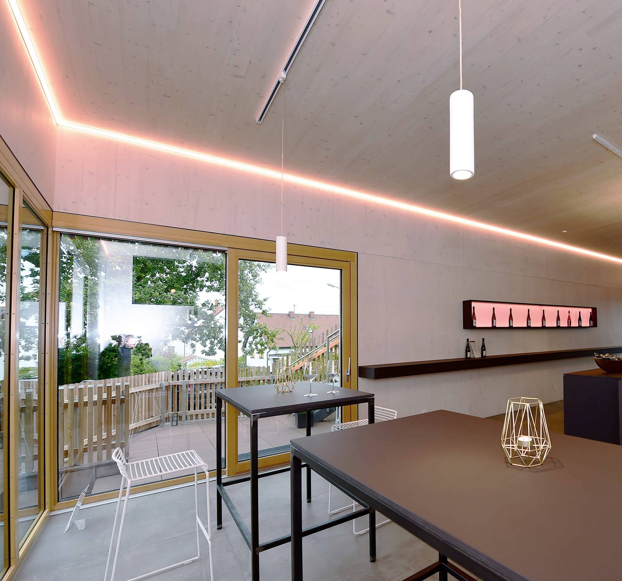 Ein modernes Café mit großen Fenstern, Holztischen und Hockern. Warmes Licht betont die Decke und eine Flasche steht auf dem Regal.