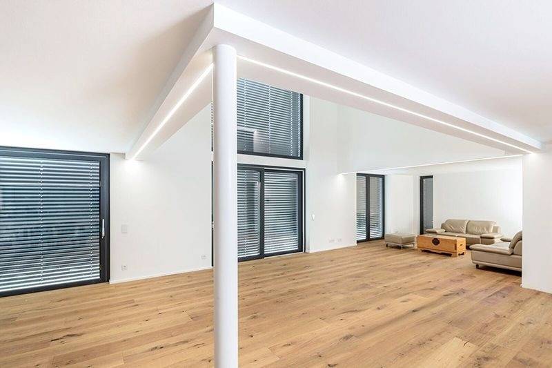 Modernes Wohnzimmer mit Holzboden, weißen Wänden und großen Fenstern mit Jalousien. Eine beige Couch steht in der rechten Ecke.