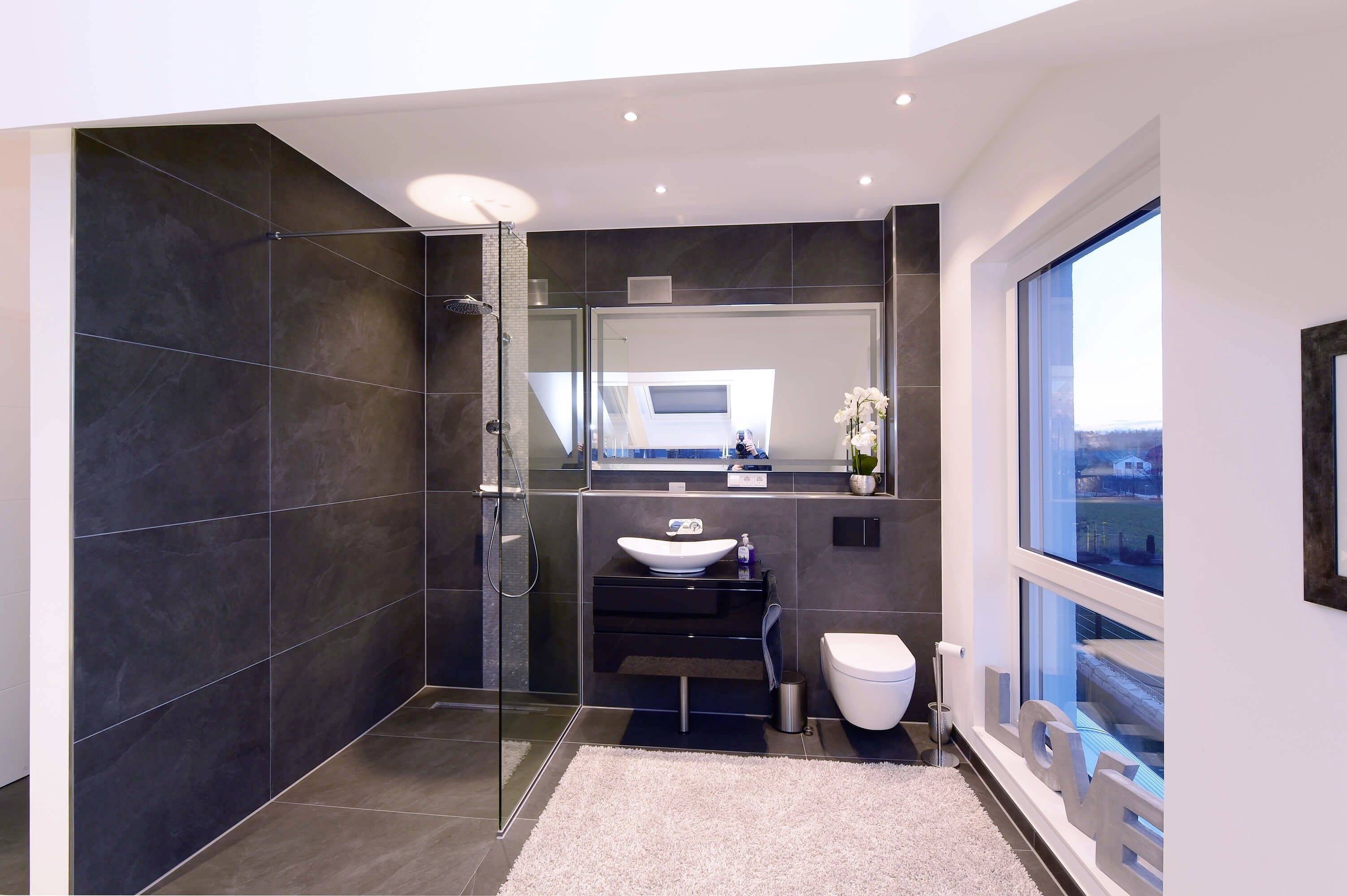 Modernes Badezimmer mit Glasduschkabine, Waschbecken, WC und großem Fenster. Helle Wände und dunkle Fliesen kontrastieren stilvoll.