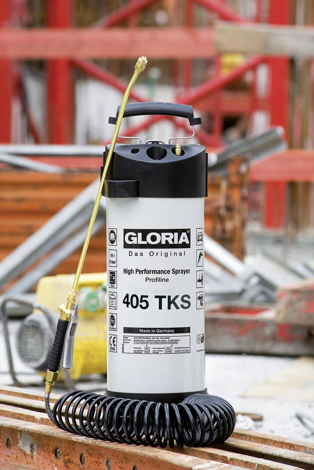 Ein Hochleistungssprühgerät mit der Aufschrift 'Gloria 405 TKS' steht vor einer Baustelle. Es hat einen goldfarbenen Sprühstab und eine Spiralschlaufe.
