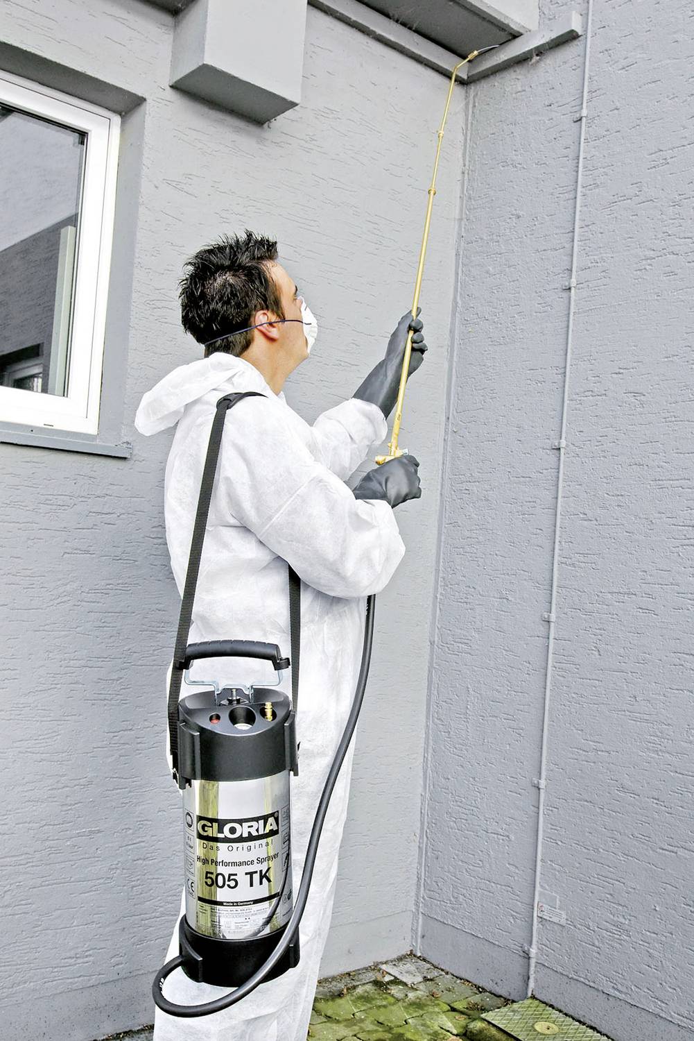 Eine Person in Schutzanzug und Maske sprüht mit einem Gloria 505 TK Drucksprüher eine Wand. Safety-Maßnahmen sind offenbar im Fokus.