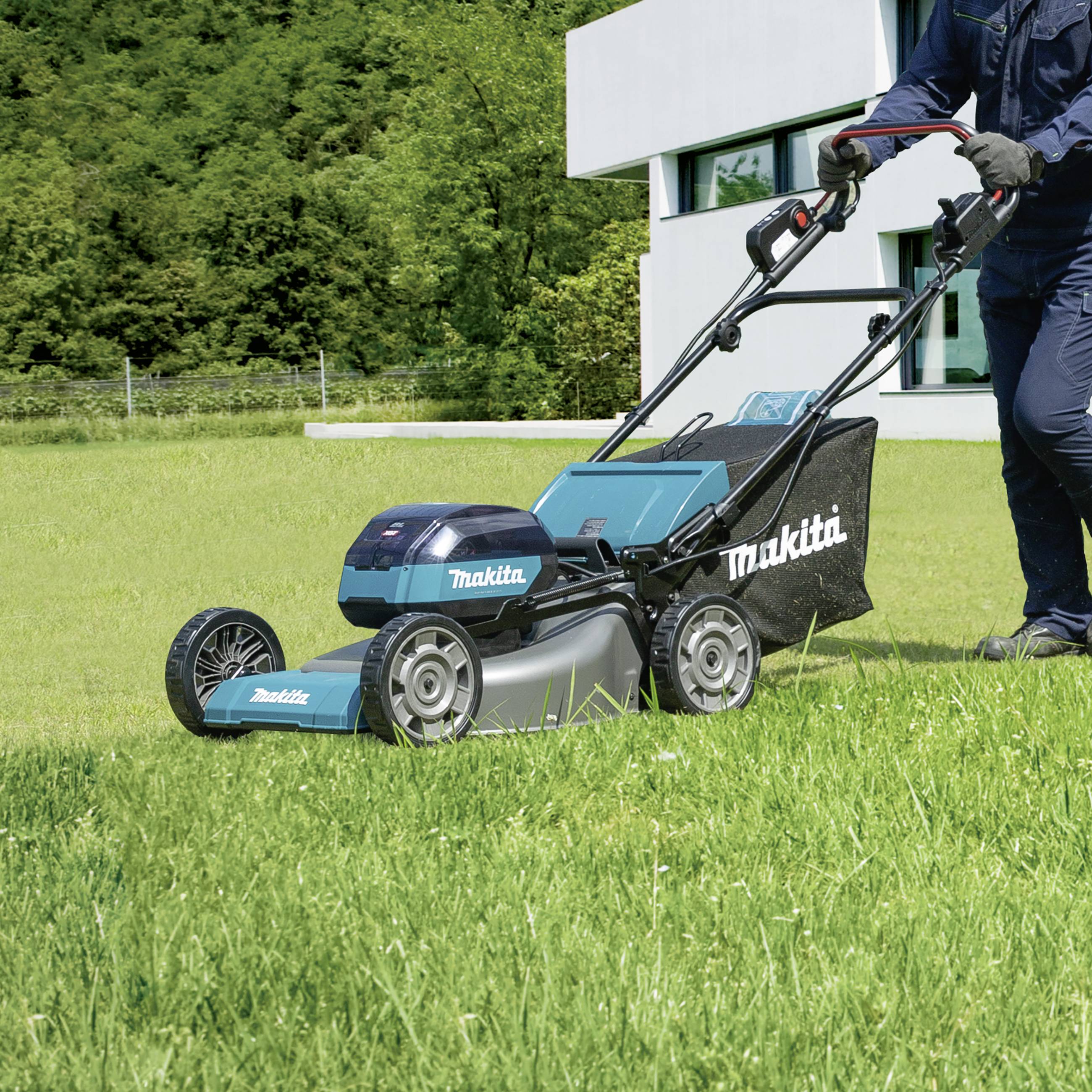 Eine Person mäht den Rasen vor einem modernen Gebäude mit einem Makita-Rasenmäher. Im Hintergrund sind Bäume zu sehen.