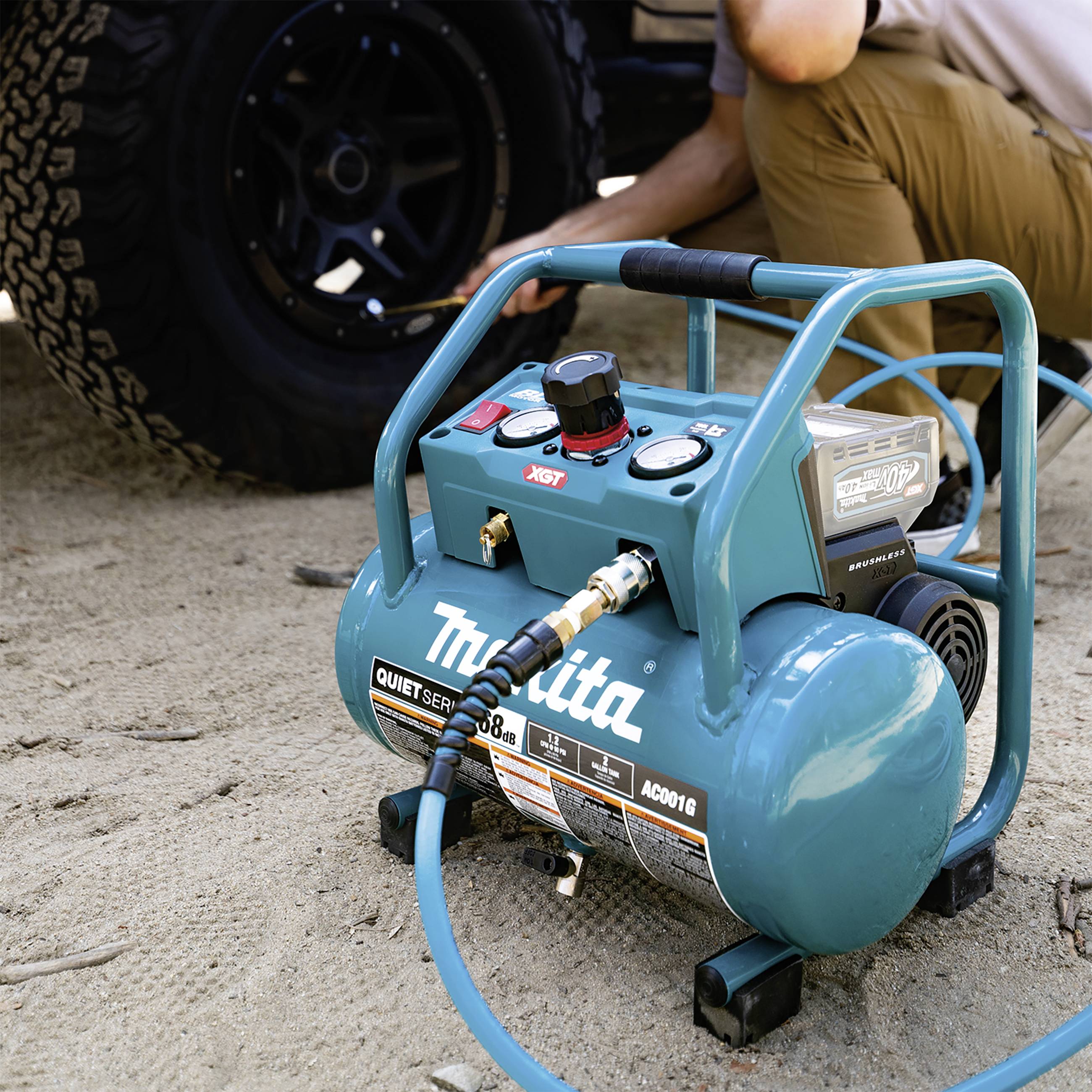 Ein tragbarer Luftkompressor der Marke Makita wird von einer Person neben einem Geländewagen in einem sandigen Umfeld benutzt.