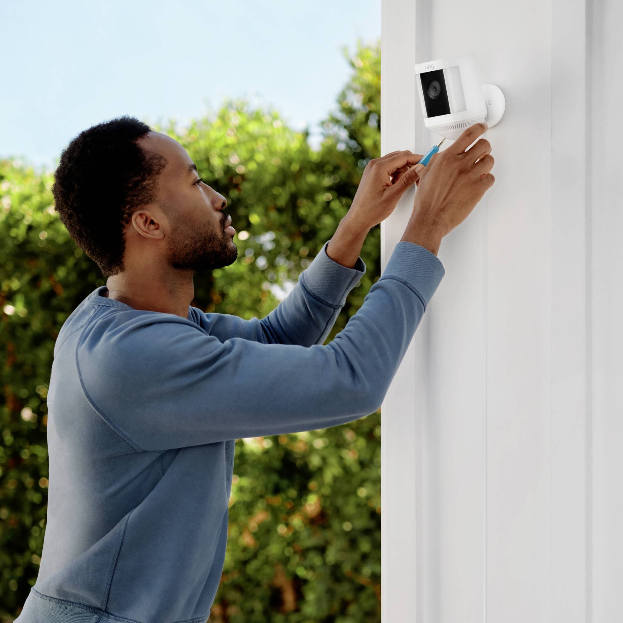 Ein Mann installiert oder justiert eine Überwachungskamera an einer Außenwand. Im Hintergrund ist eine grüne Hecke zu sehen.