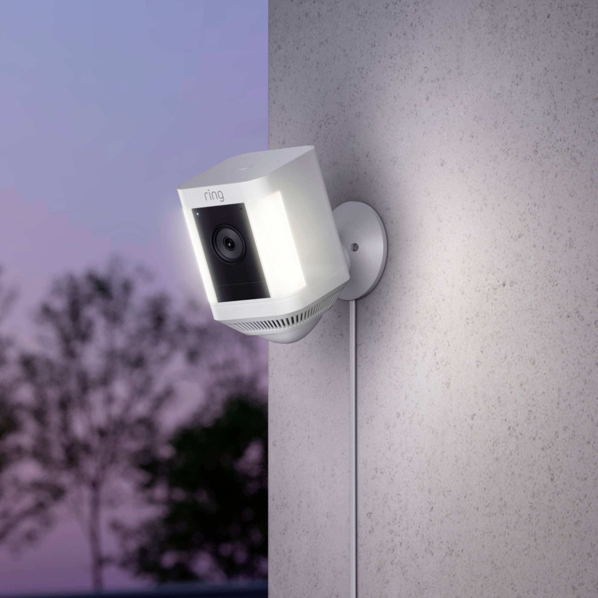 Eine Überwachungskamera mit integriertem Licht montiert an einer Wand im Freien; im Hintergrund verschwommene Bäume und Abendhimmel.