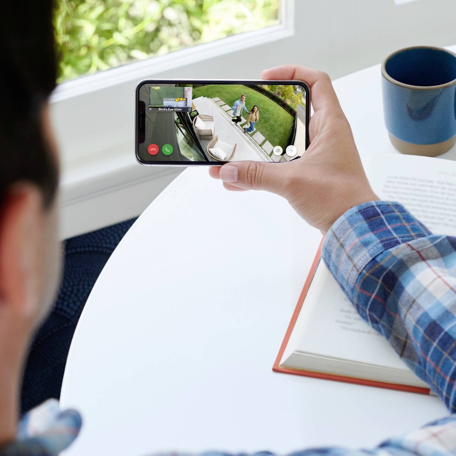 Eine Person sieht ein Video auf einem Smartphone an, das einen Lieferdienst auf einer Veranda zeigt. Im Hintergrund liegt ein Buch.