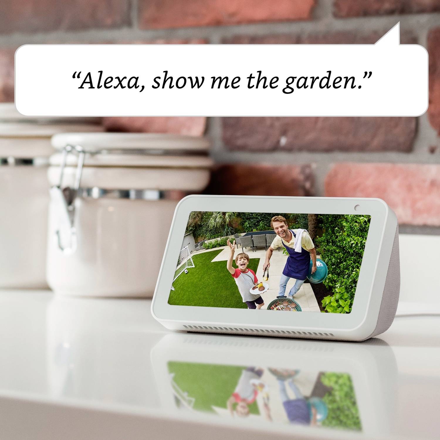 Ein Smart-Display auf einem Küchentisch zeigt ein Foto von Vater und Kind im Garten. Text in Sprechblase: 'Alexa, zeige mir den Garten.'