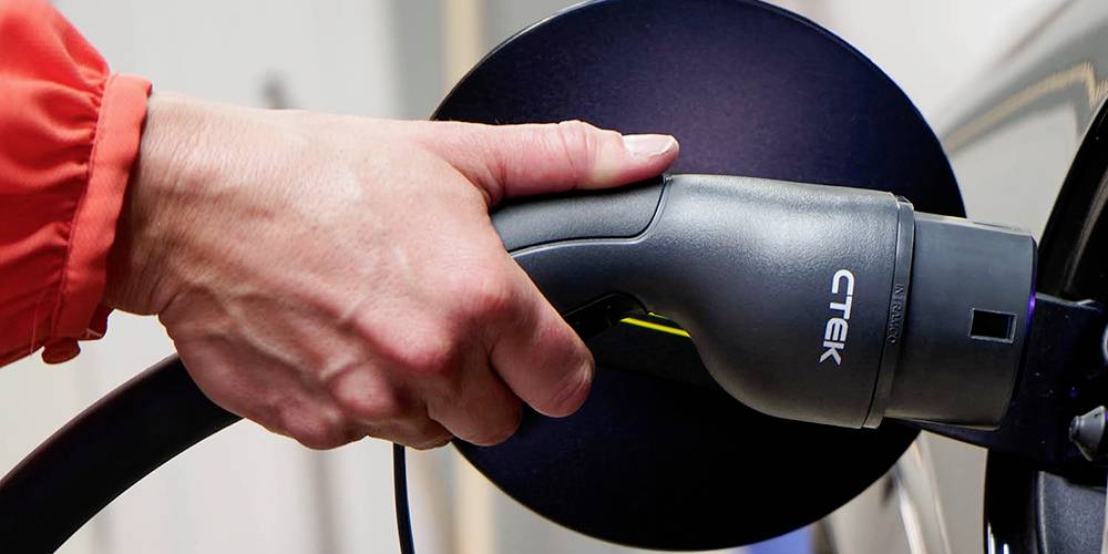 Eine Hand hält einen Elektroauto-Ladestecker und verbindet ihn mit einem Auto. Ein Aufdruck zeigt 'CTEK' auf dem Stecker.