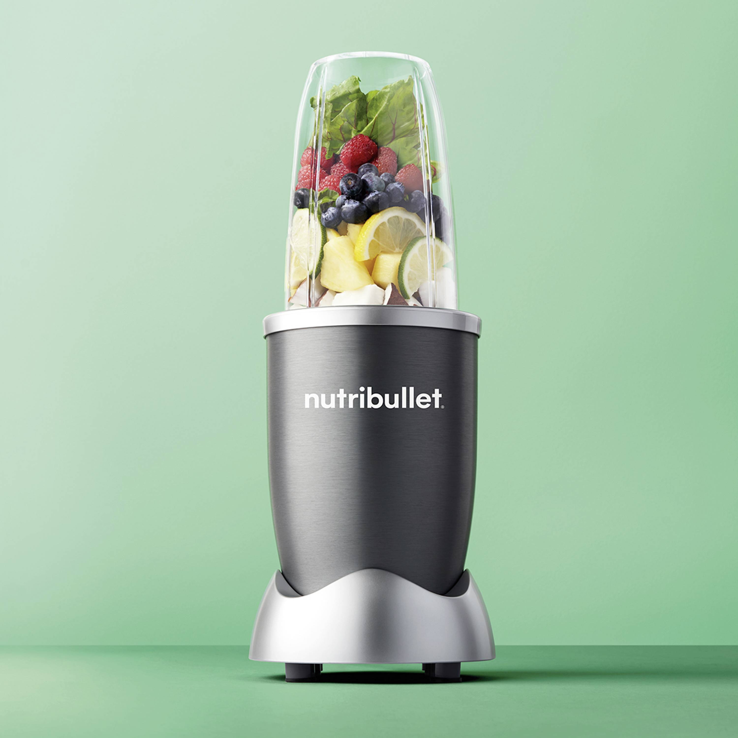 Ein grauer Mixer mit Obst und Gemüse darin, darunter Beeren und Zitronenscheiben, vor grünem Hintergrund. Text 'nutribullet' auf Gerät.