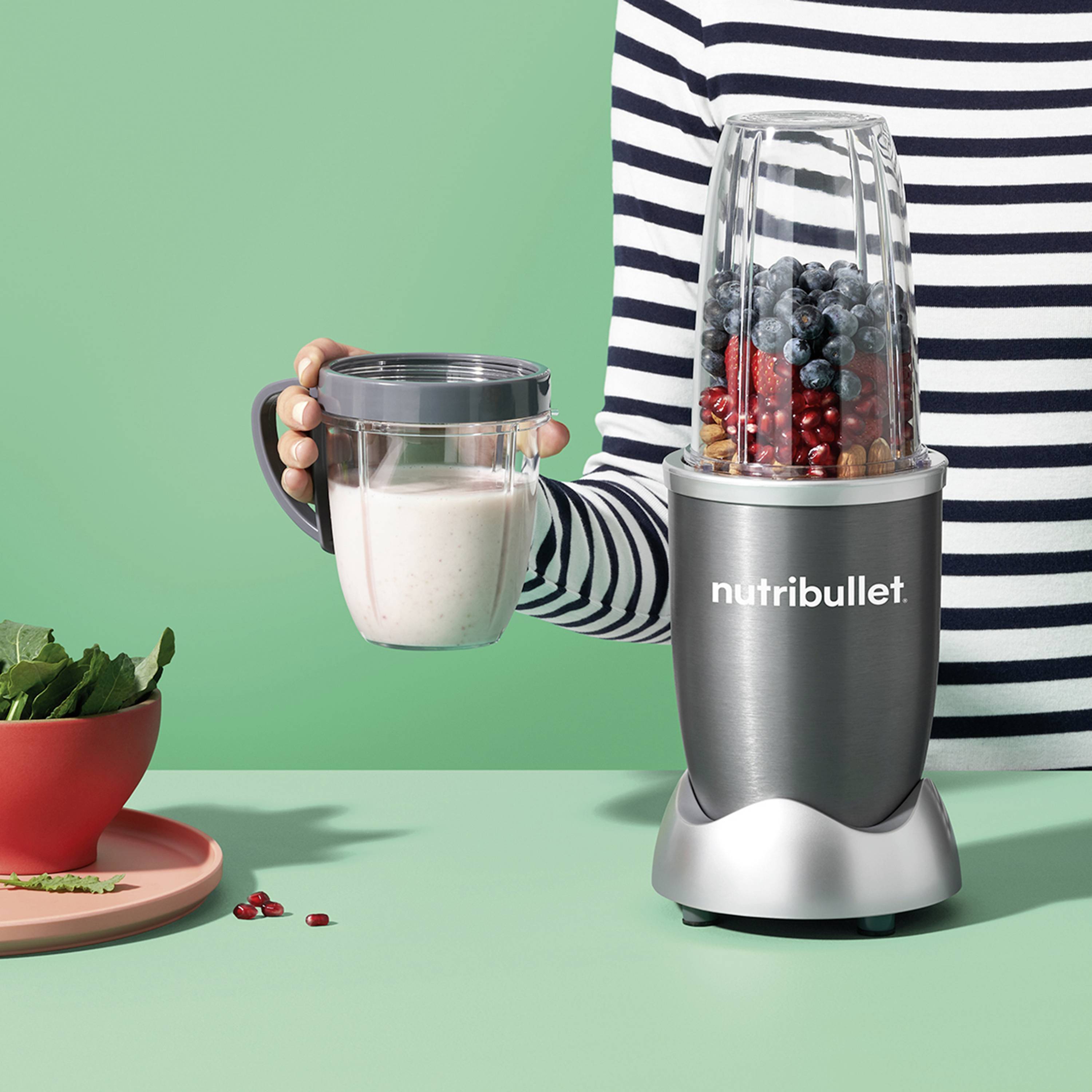 Eine Person hält einen Becher mit Schäumgetränk neben einem grauen Mixer, gefüllt mit Blaubeeren und Nüssen. Ein Teller mit Minzblättern.