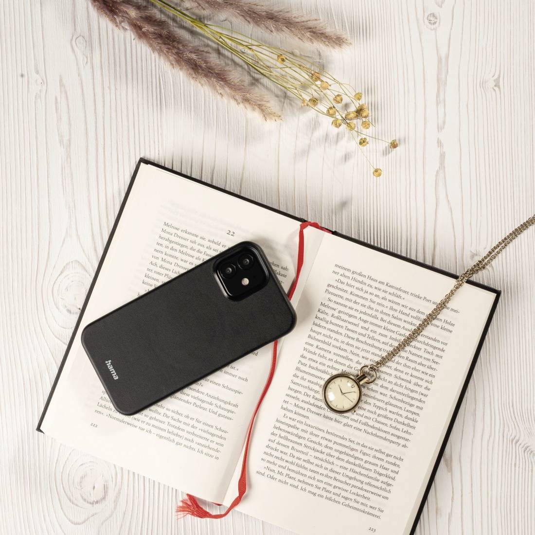 Aufgeschlagenes Buch auf Holztisch mit schwarzem Smartphone und Taschenuhr darauf platziert, dekoriert mit getrockneten Pflanzen.