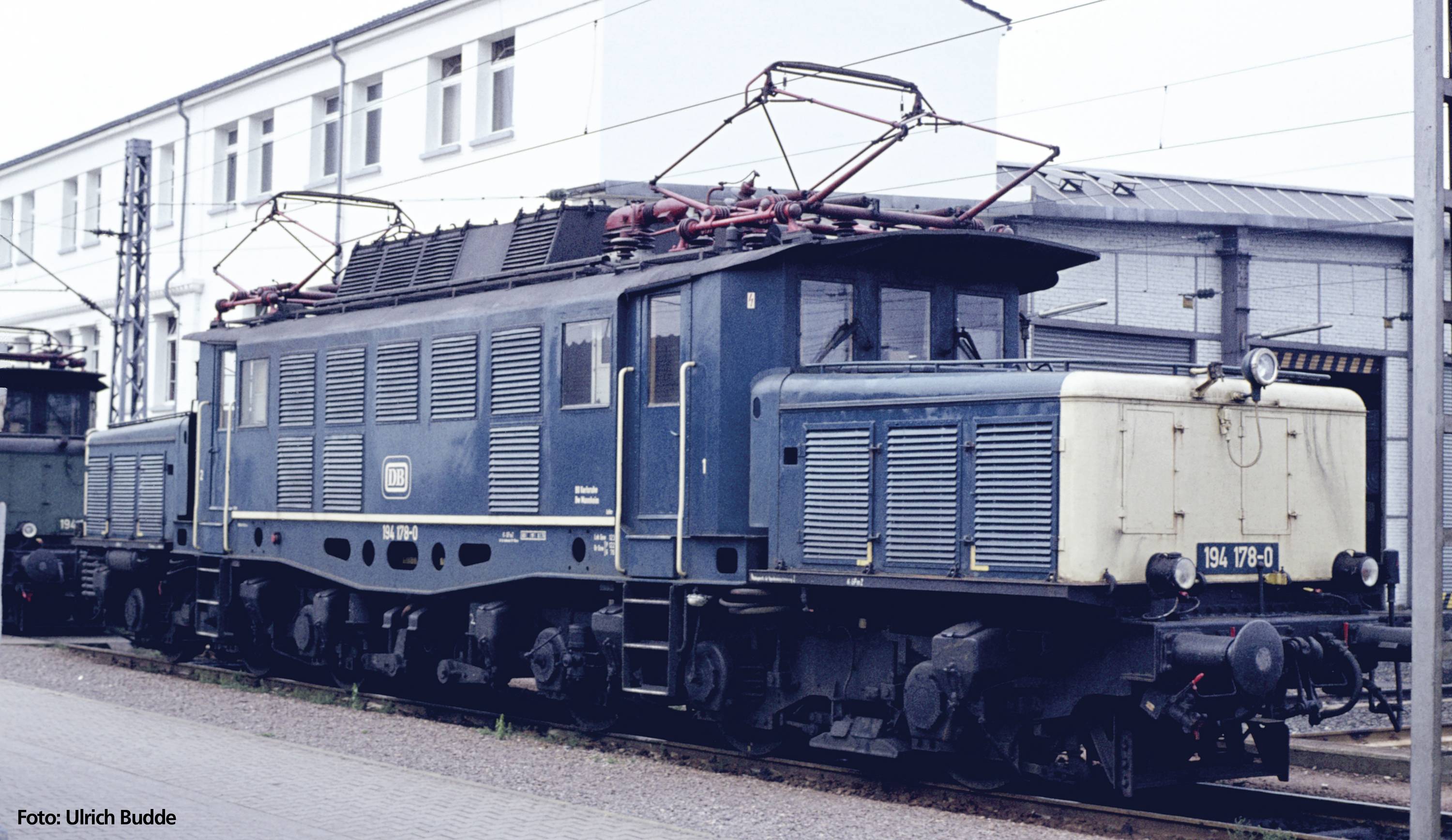 Eine blaue elektrische Lokomotive steht auf einem Bahngleis vor einem Gebäude. Sie trägt die Aufschrift 'E 94 178'.