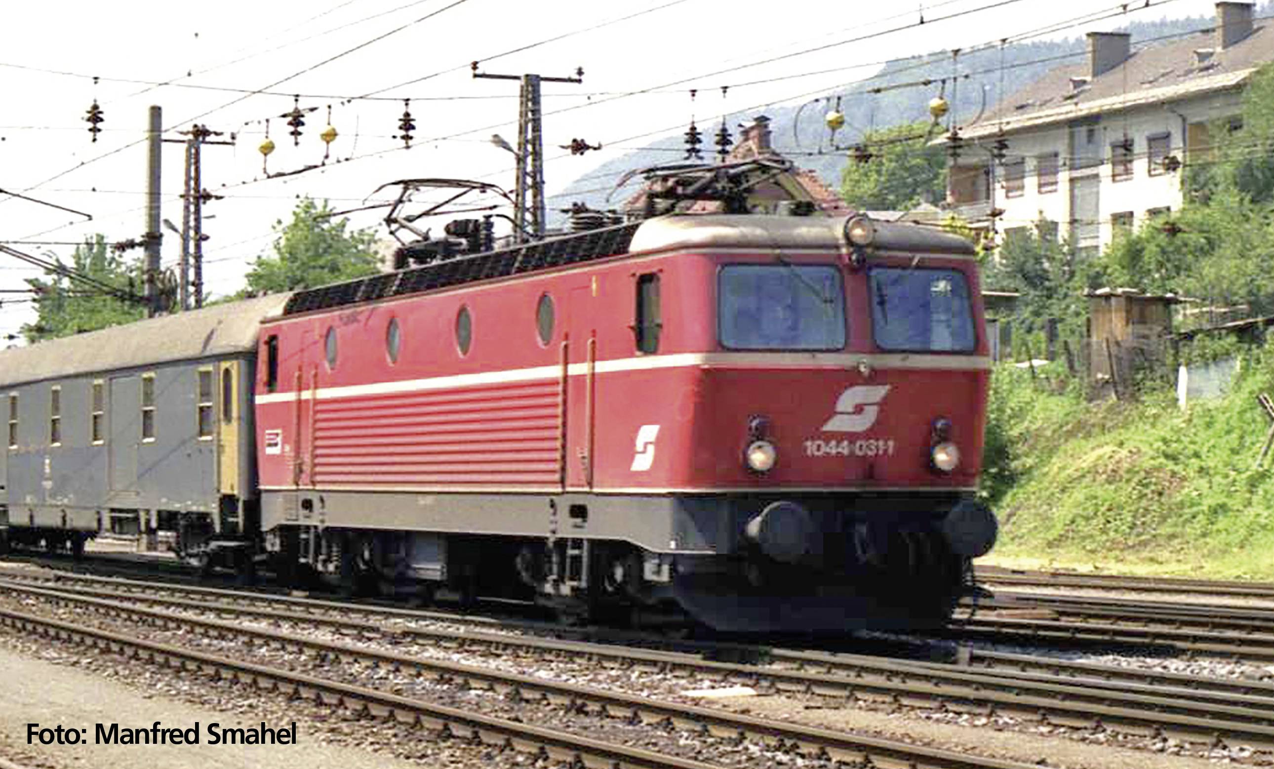 Eine rote Elektrolokomotive zieht einen Personenzug über Bahngleise, im Hintergrund sind Oberleitungen und Gebäude zu sehen.