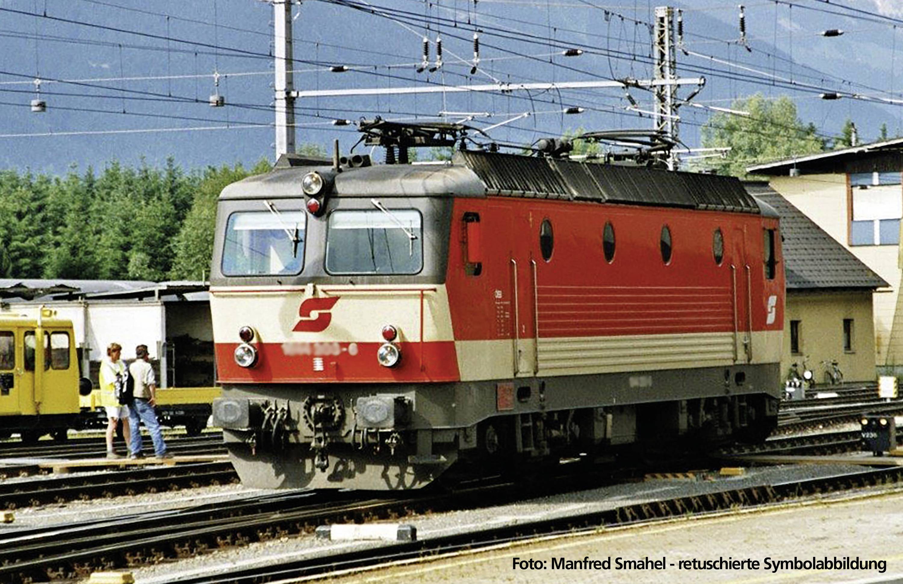 Eine rote und graue elektrische Lokomotive steht auf einem Bahnhofsgleis, umgeben von anderen Bahngleisen und Gebäuden im Hintergrund.