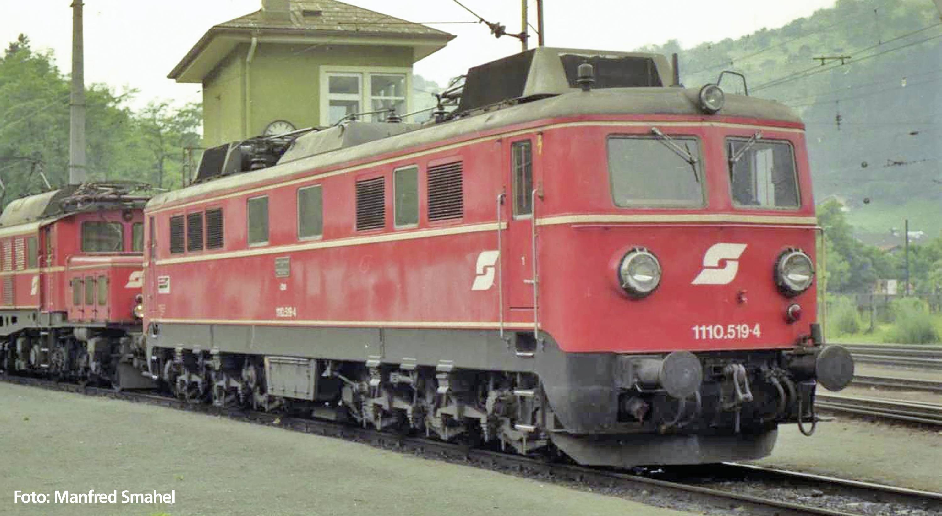 Rote Elektrolokomotive der Baureihe 1110 mit grauem Dach steht auf einem Gleis vor einem Bahnwärterhäuschen. Waldige Hügel im Hintergrund.