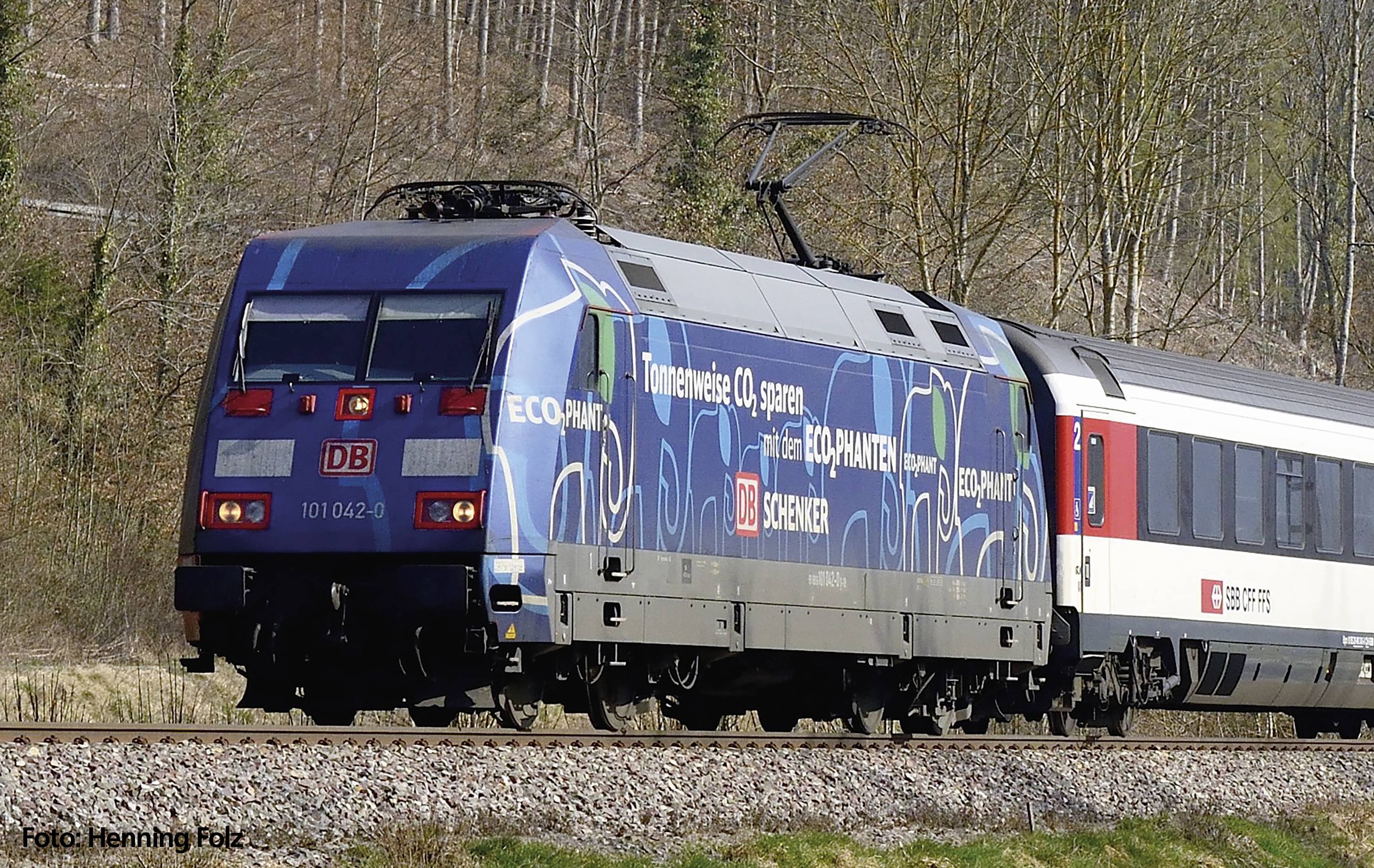 Ein blauer Elektrozug der Deutschen Bahn fährt auf einem Gleis durch eine Waldlandschaft. Aufschrift: 'Umwelt schützen'.