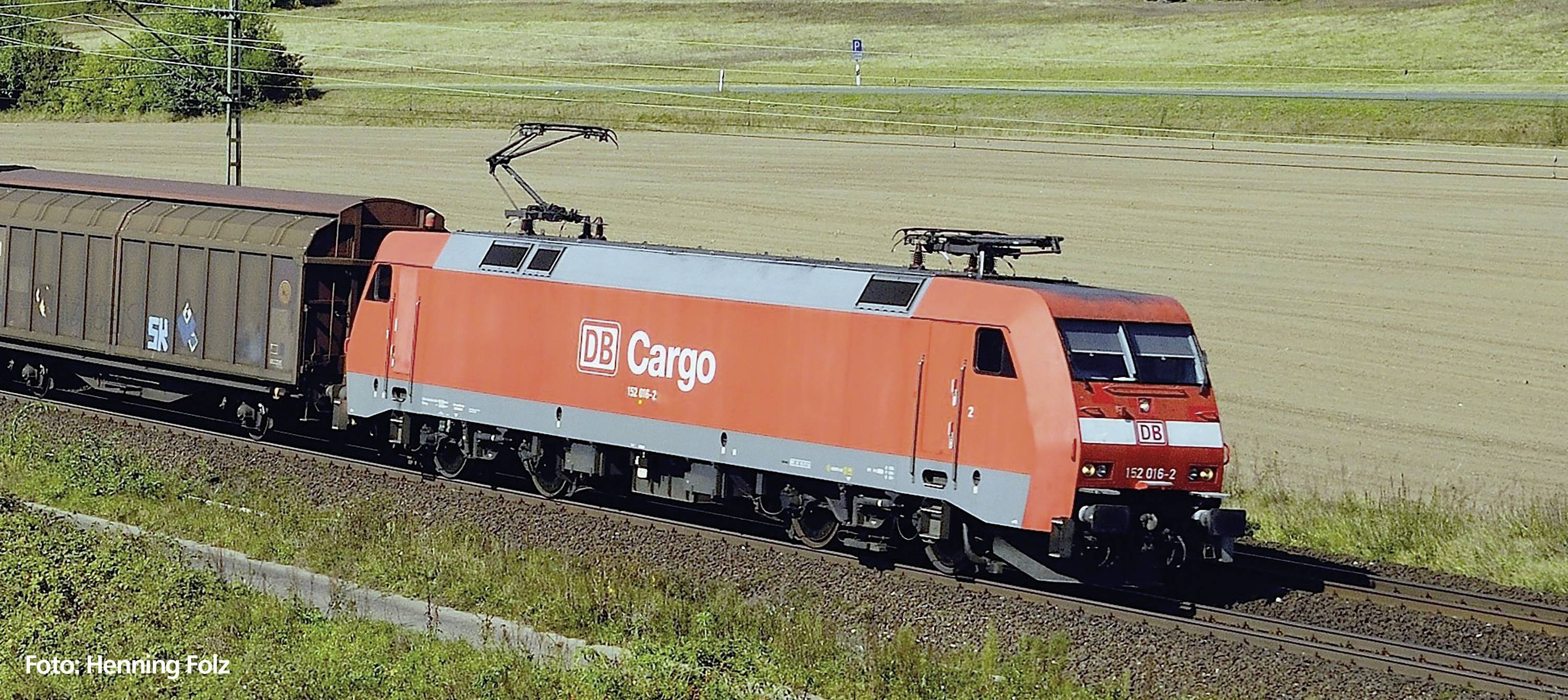 Ein roter Güterzug der DB Cargo fährt auf Schienen durch ländliche Umgebung bei Tageslicht. Links im Bild befindet sich ein Güterwaggon.