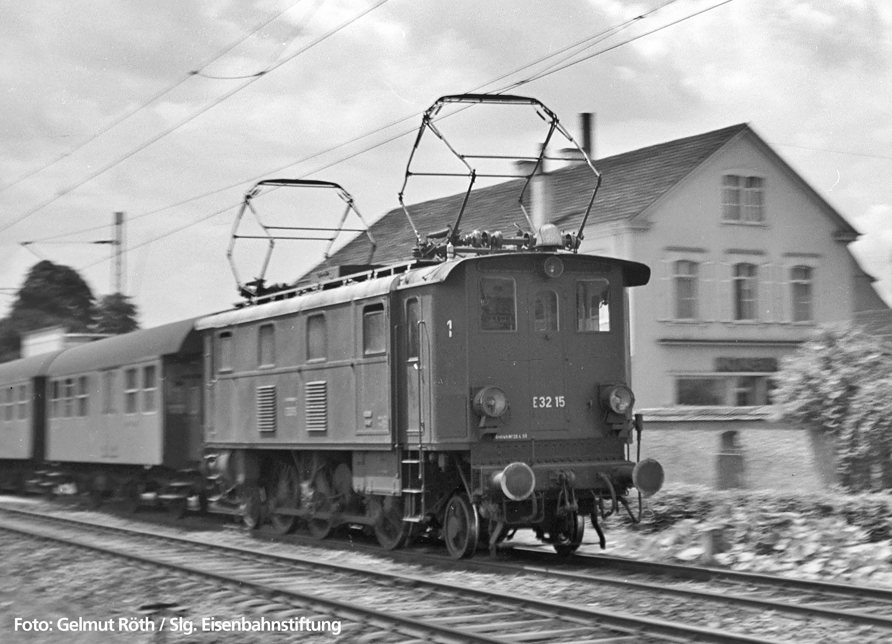 Schwarz-weiß Foto einer historischen Elektrolokomotive E 32 15 fährt vor einem Passagierwaggon an einem Gebäude vorbei.