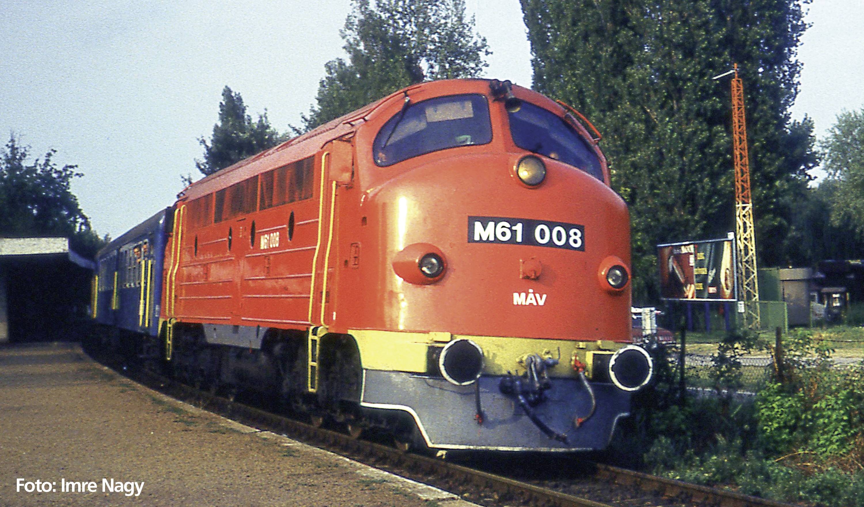 Rote Diesellokomotive M61 008 der MÁV auf einem Bahngleis, umgeben von Bäumen. Links ist ein kleiner überdachter Bereich zu sehen.