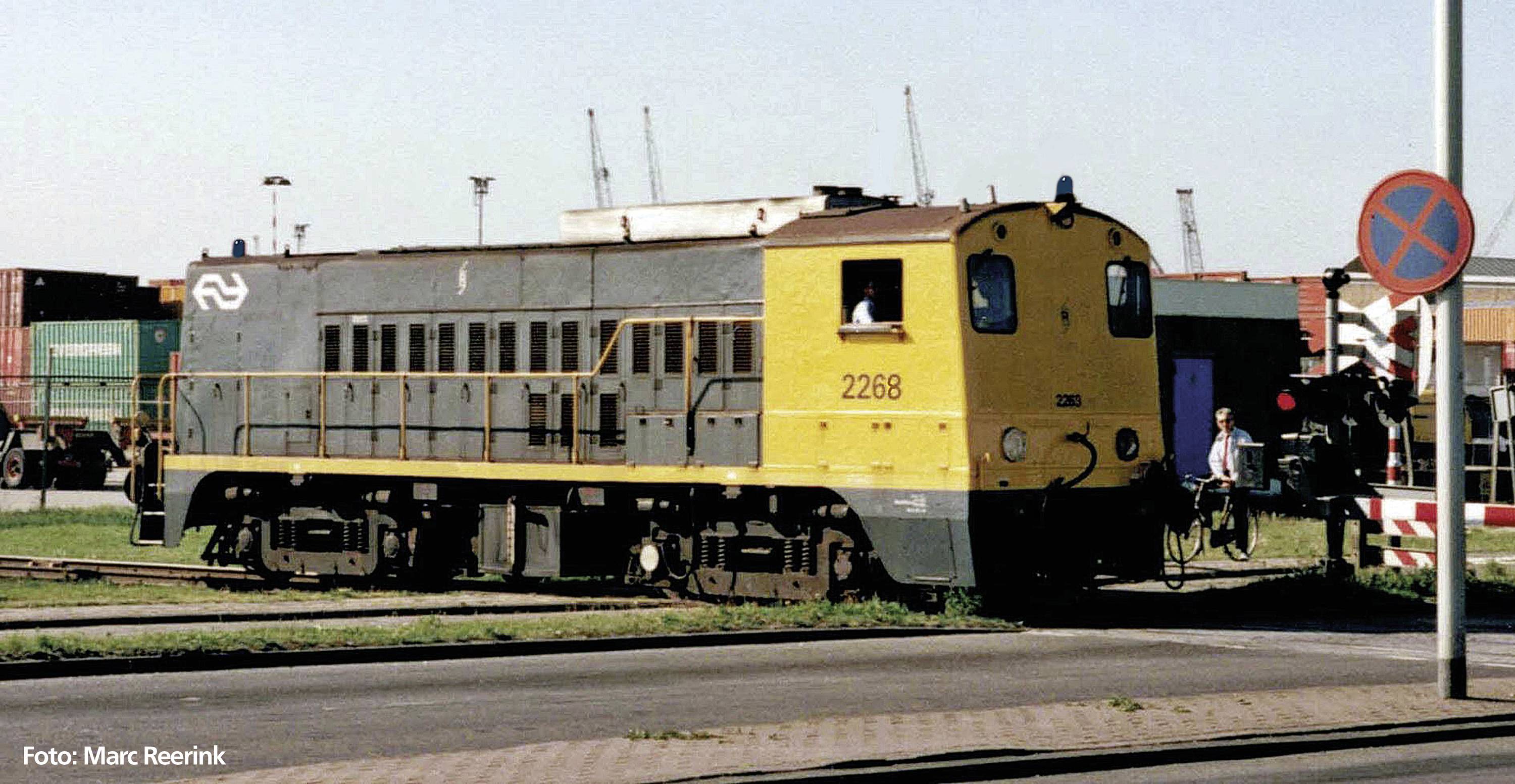 Eine gelbe und graue Lokomotive mit der Nummer 2268 fährt auf einem Gleis in einem Industriegebiet. Ein Zebrastreifen und ein Schild sind sichtbar.