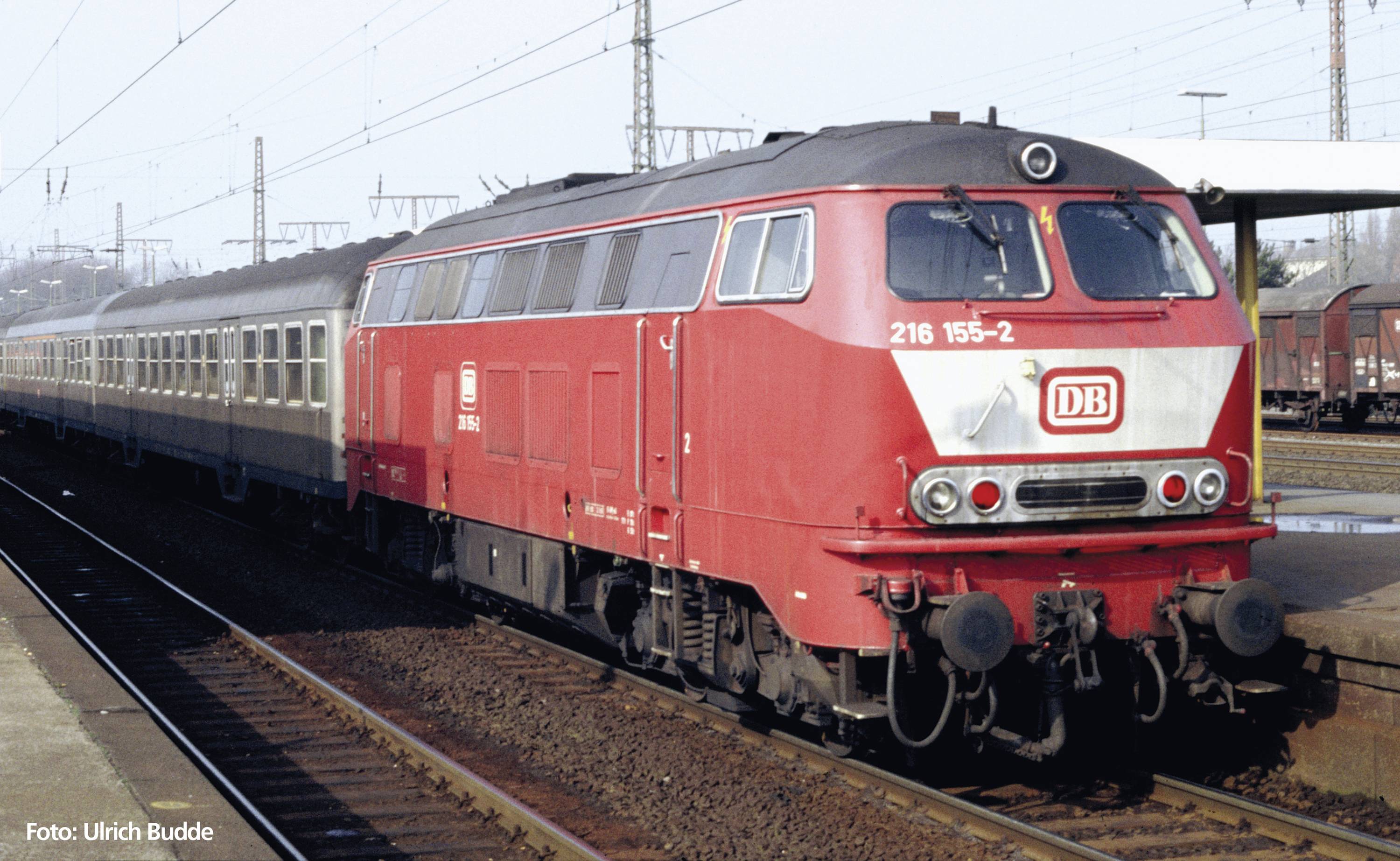 Roter DB-Zug mit Diesellokomotive 216 155-2 zieht Personenwagen auf einem Bahngleis bei Tageslicht. Schienen und Oberleitungen sind sichtbar.