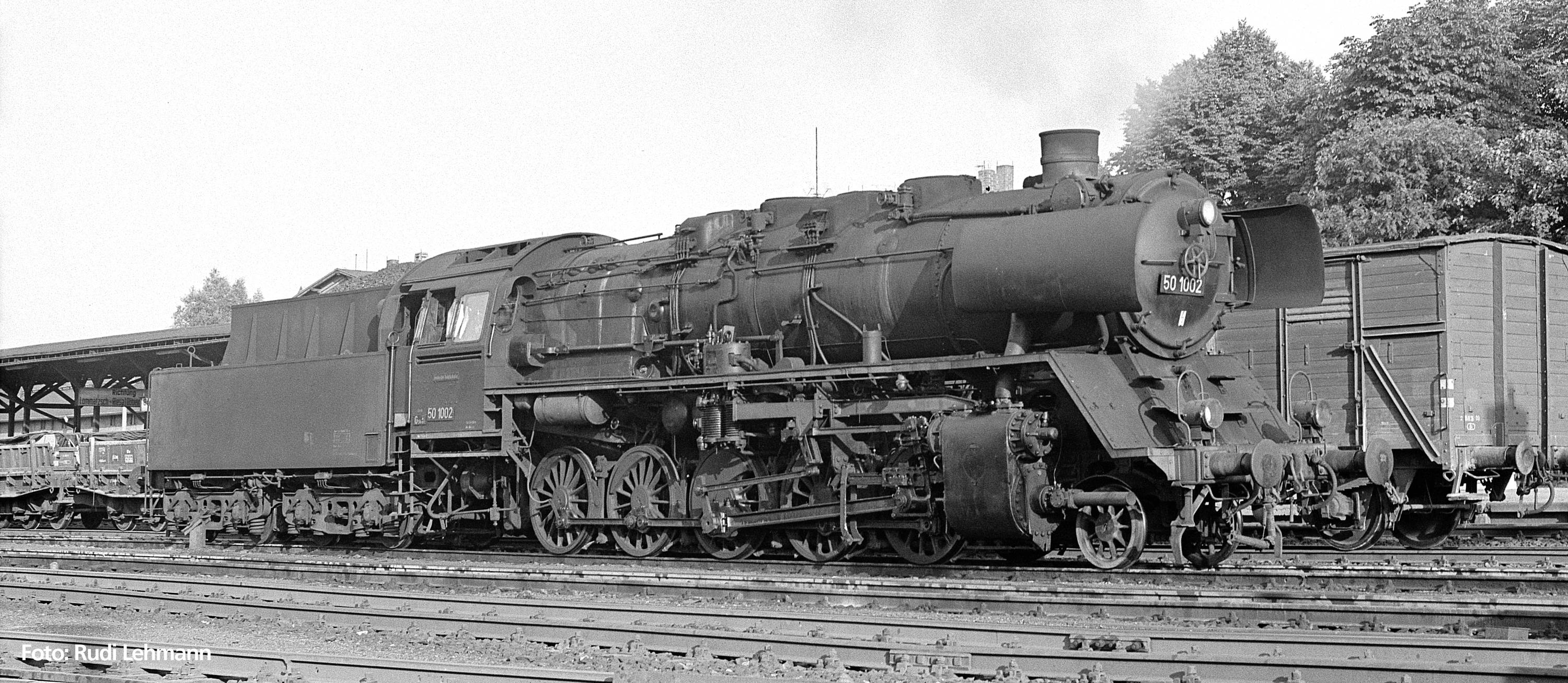 Eine Dampflokomotive steht auf einem Bahngleis. Im Hintergrund sind ein Bahnhof und Bäume sichtbar. Die Lokomotive ist groß und massiv.